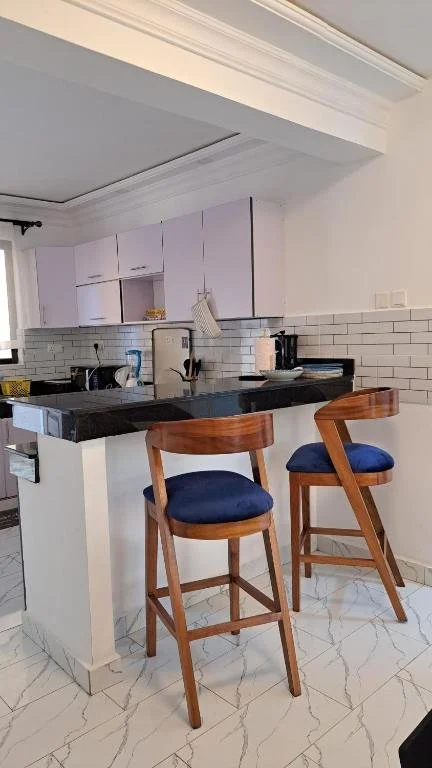 Modern kitchen with white cabinets, black countertop, white subway tile backsplash, two wooden bar stools with blue cushions, and visible appliances and decor.
