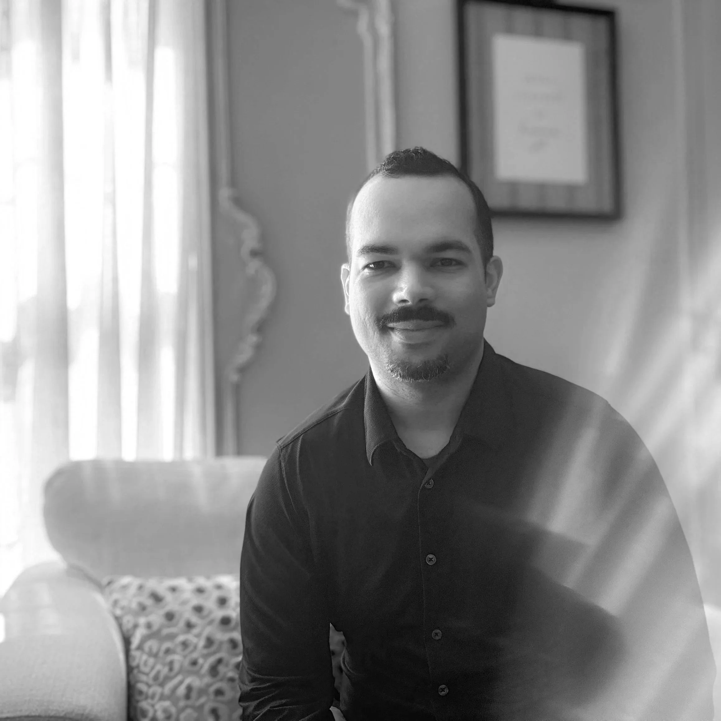A young man with short hair, a mustache, and a goatee smiling in a black button-up shirt, standing indoors with a patterned carpet, white curtains, and framed artwork on the wall behind him.