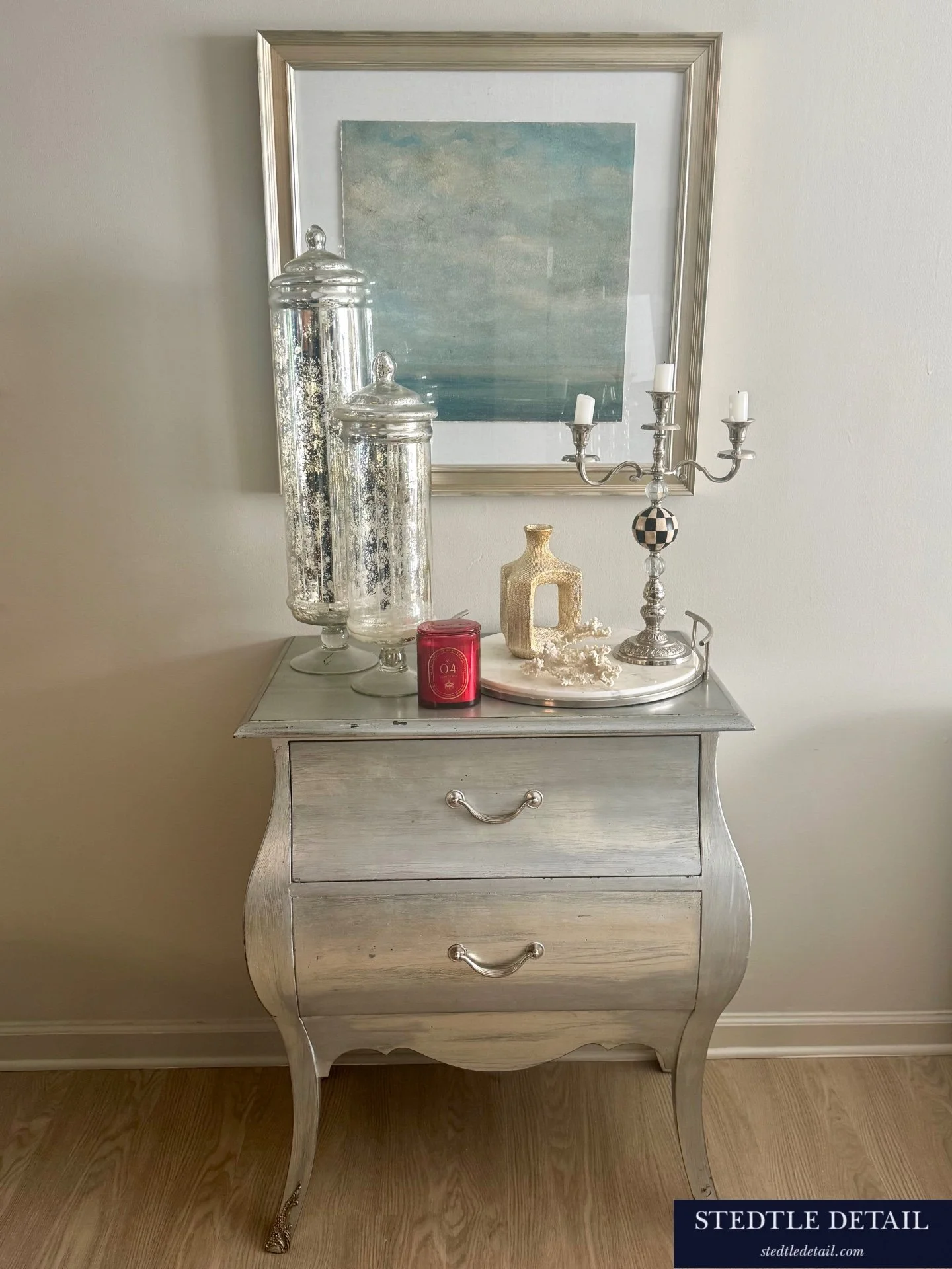 A white vintage dresser with three drawers and ornate curved legs, topped with decorative items including tall glass jars, a small cream-colored vase, white coral, a red candle, and a silver candelabra with three unlit candles. A framed watercolor painting of a seascape hangs on a plain white wall above the dresser.