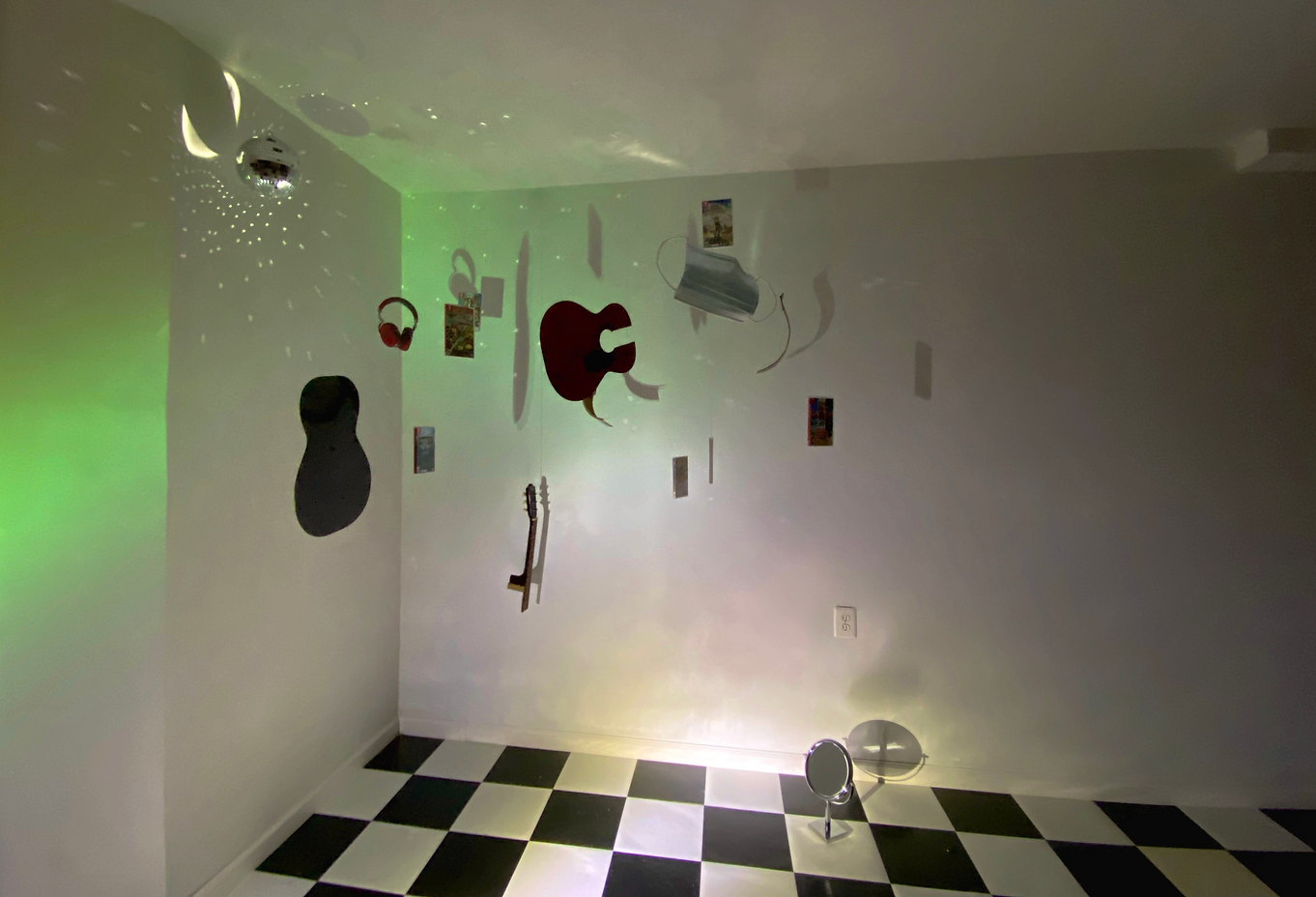 Room with black and white checkered floor, white walls, decorated with hanging photographs, cutouts of a guitar, headphones, and abstract shapes, and illuminated with colorful lights and reflections.