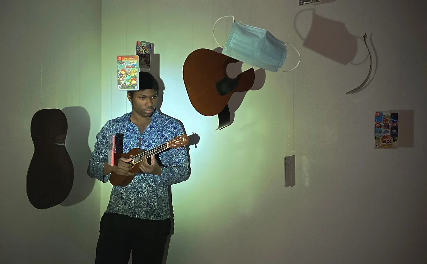 A young man holding a ukulele, standing against a wall with various objects hanging, including a face mask, a guitar, and posters.