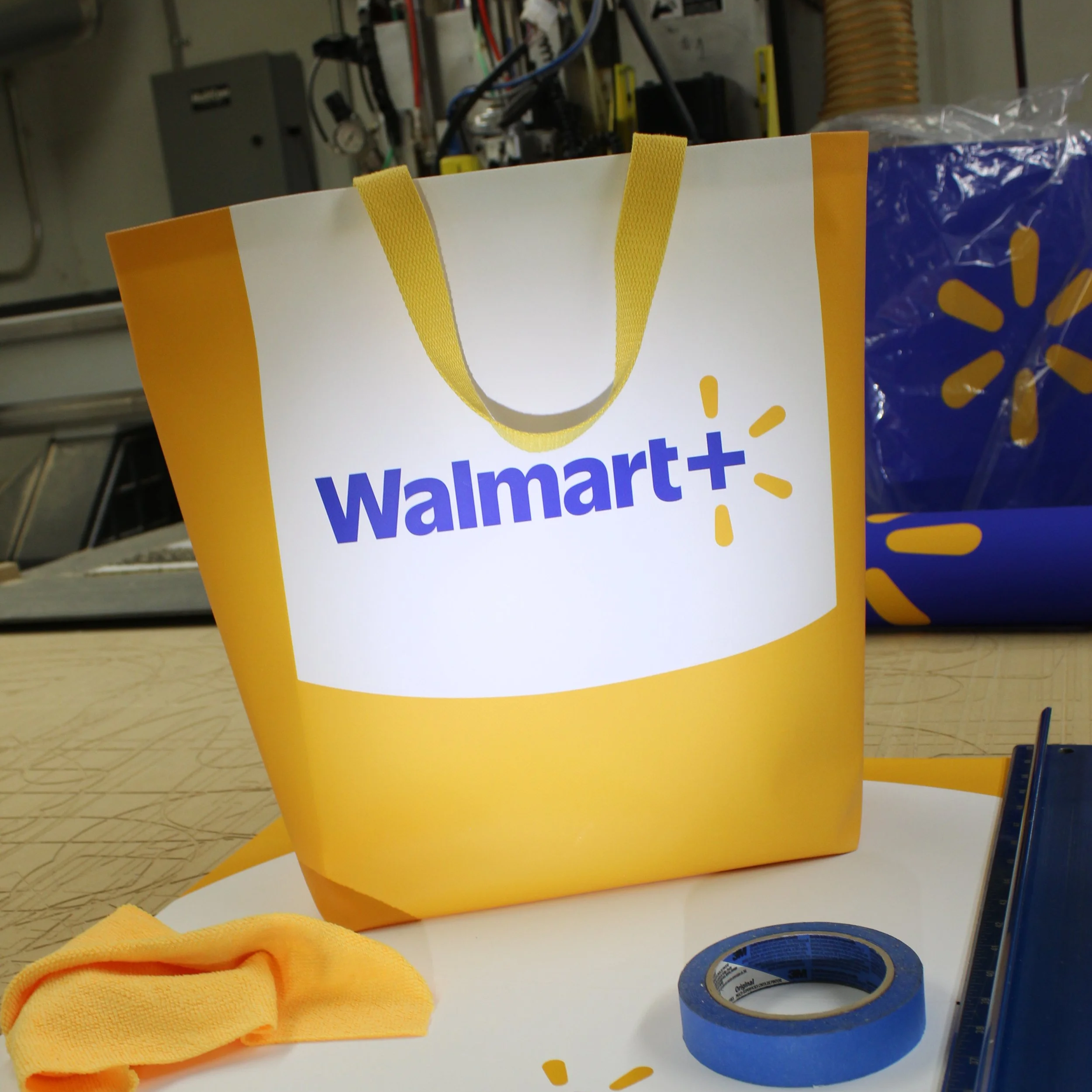 A Walmart shopping bag with yellow handles and branding on the front, placed on a table with a roll of blue painter's tape and a yellow cloth or clothing item nearby. In the background, there are various objects and equipment, including control panel