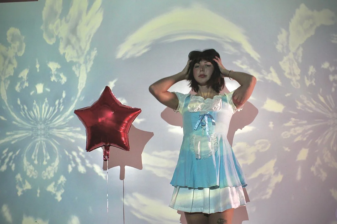 A woman wearing a pastel dress standing against a background with cloud and star patterns projected on the wall. She is holding her head with both hands and next to her is a red star-shaped balloon.