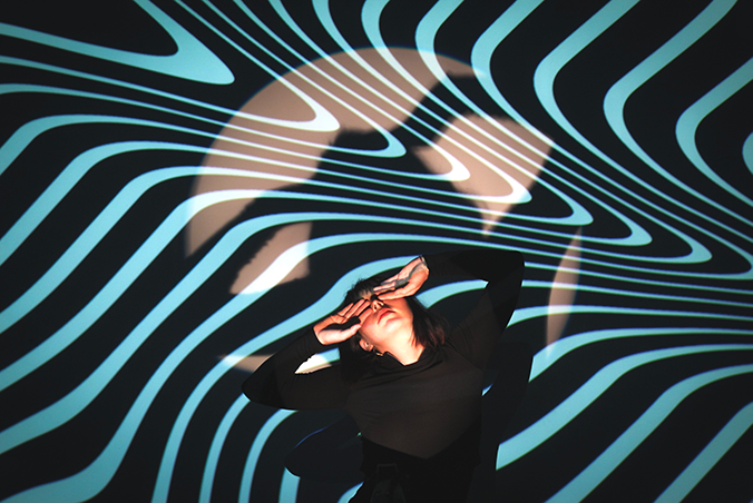 Person standing in a dark room with blue and white light projections creating swirling patterns on the walls and ceiling.
