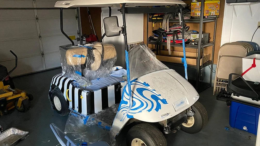 A golf cart with a decorated windshield and seats covered in plastic, parked inside a garage surrounded by various tools and equipment.