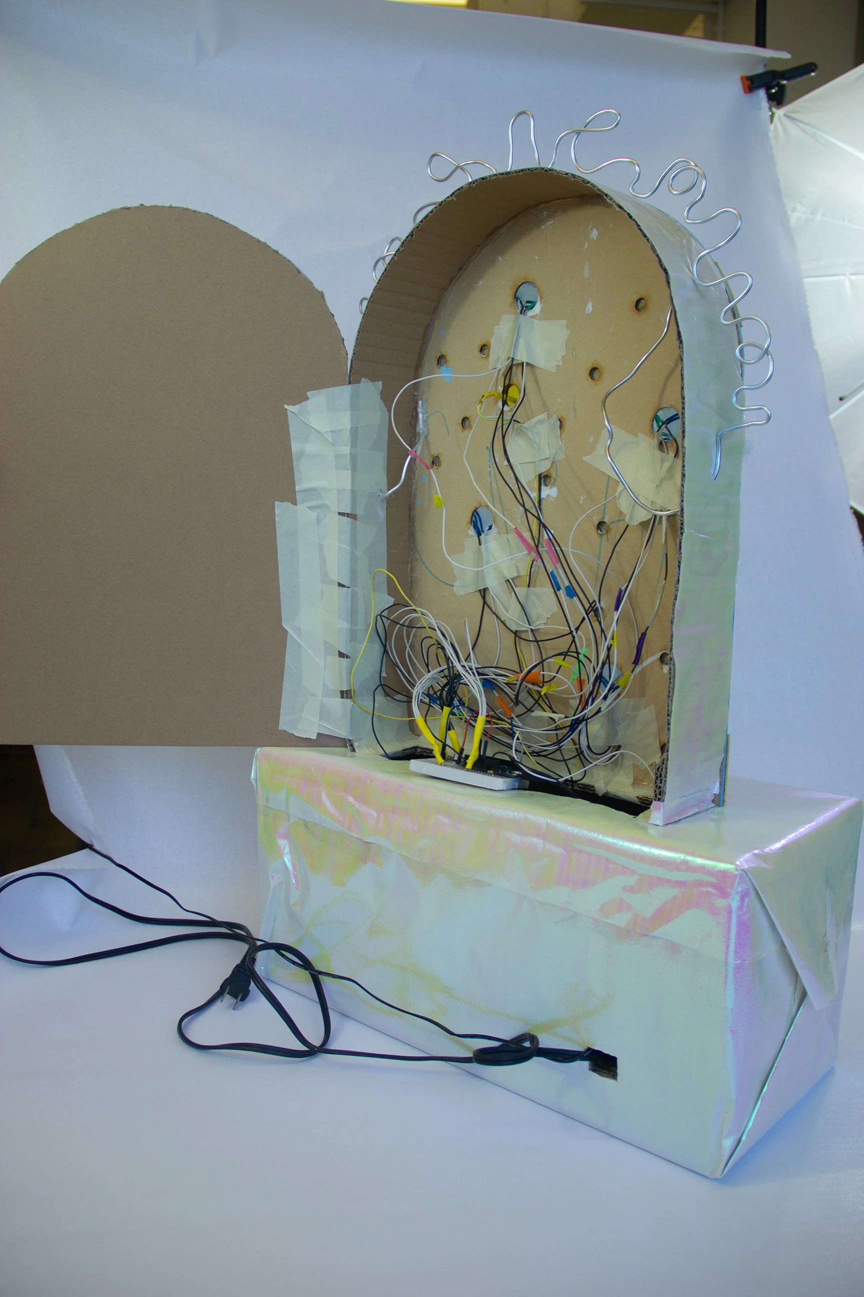 Close-up of a cardboard moon-shaped object with wires and electronic components inside, covered with iridescent wrapping paper, on a white table.