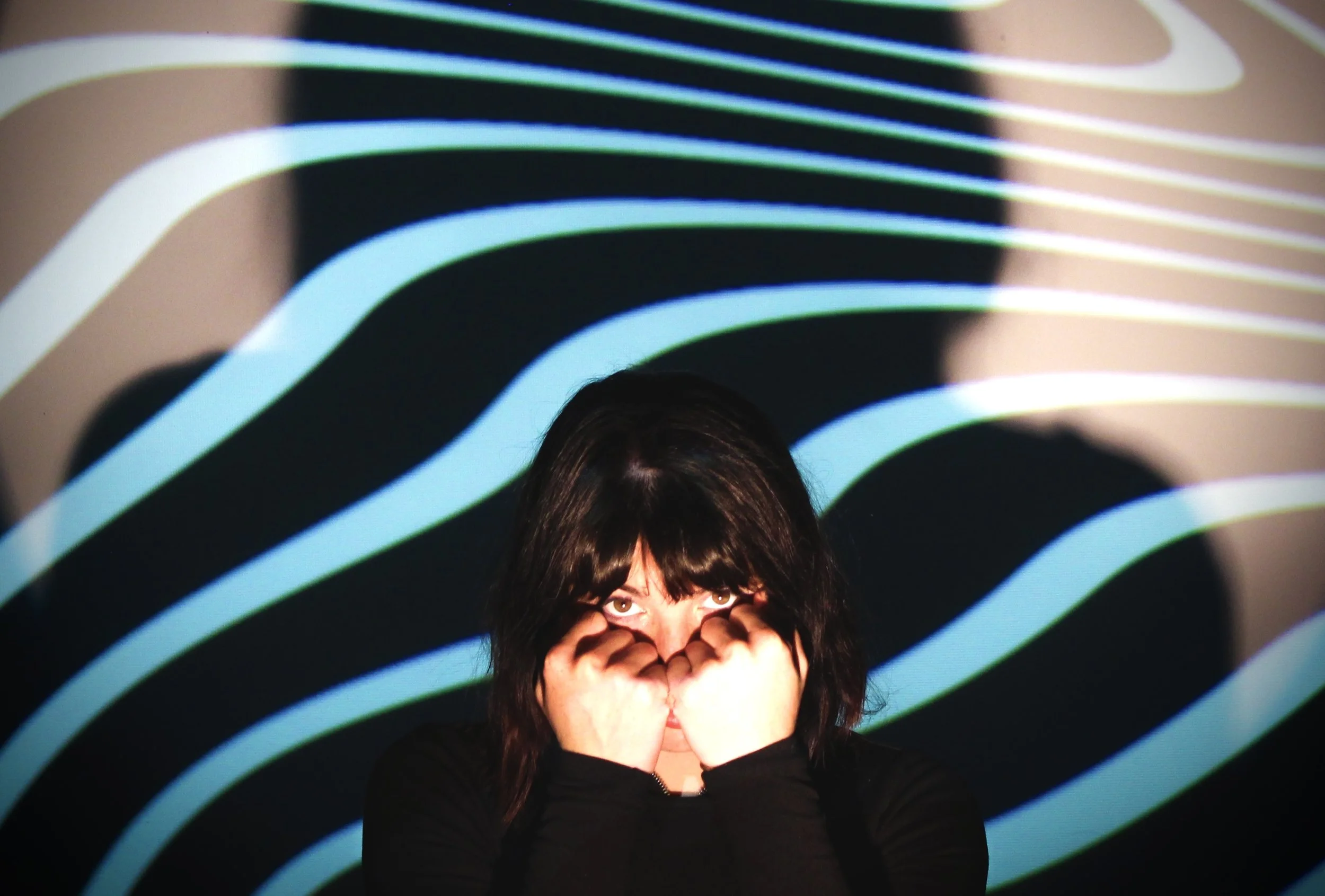 A person with dark hair, wearing a black top, covers their face with their hands. They are standing in front of a black background with colorful wavy lines projected onto it, creating a vibrant, abstract pattern.