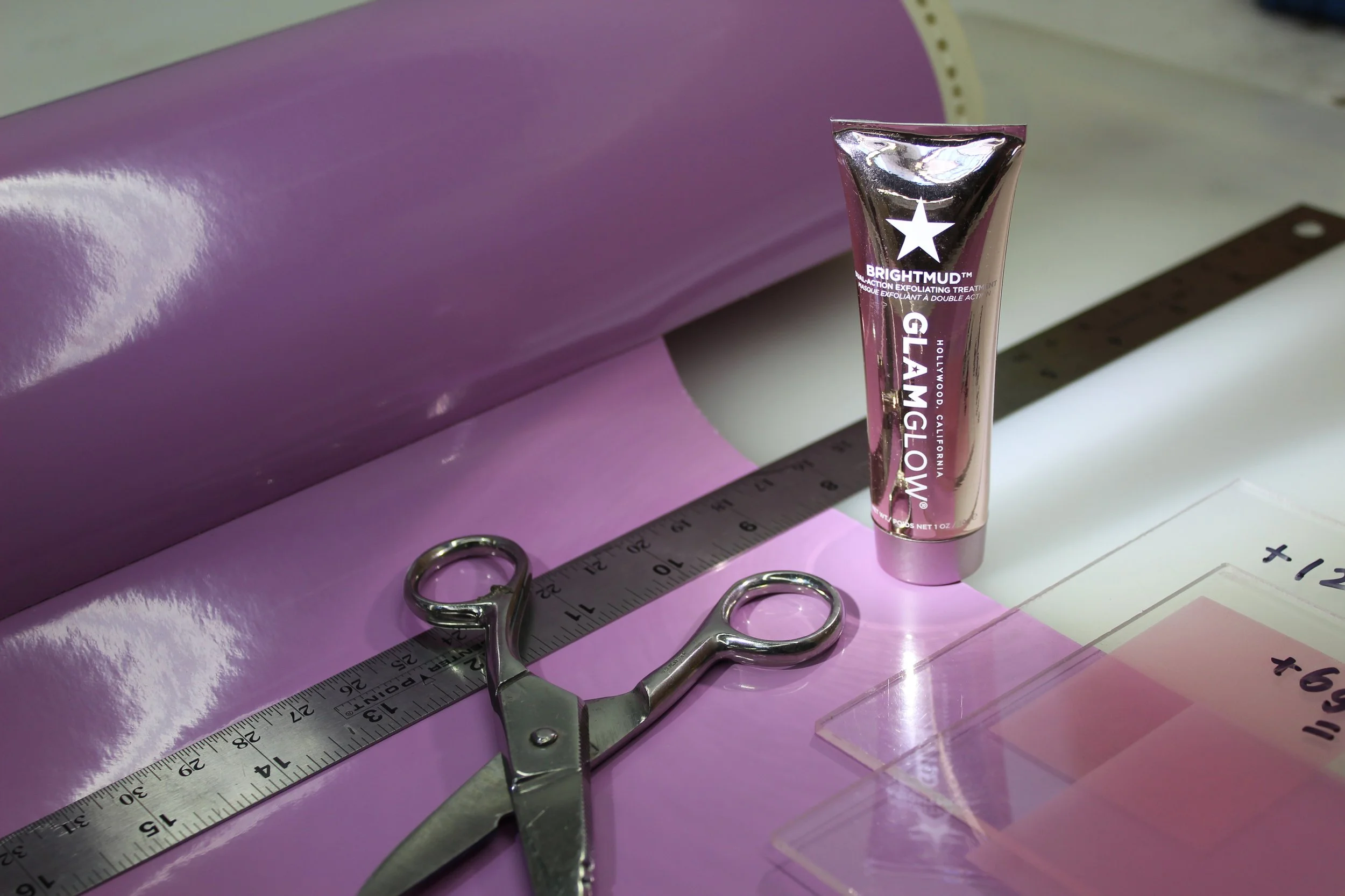 A tube of Brightmud GlamGlow exfoliating treatment stands upright on a pink surface, with a metal ruler, a pair of scissors, and a small clear plastic container nearby. The background includes a purple object and white paper.