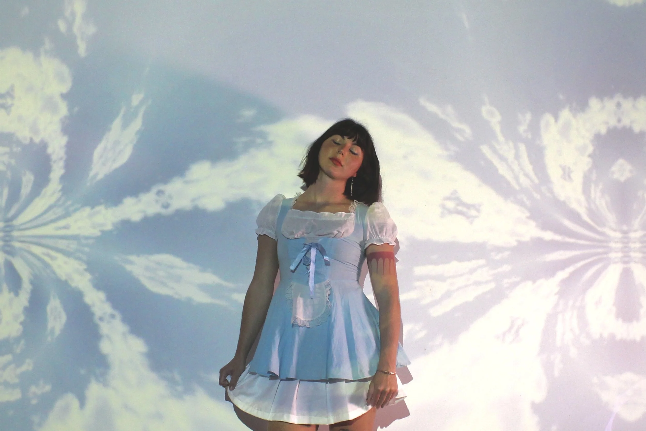 A young woman with dark hair and bangs wearing a white dress with puffed sleeves and a blue ribbon, standing with a projected view of clouds behind her.