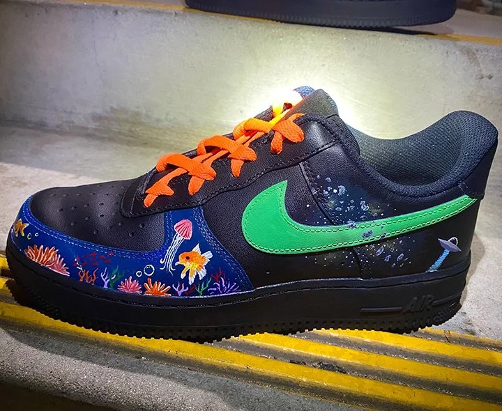 A customized black Nike Air Force 1 sneaker with a green swoosh, orange laces, and a hand-painted underwater scene featuring fish, coral, and a jellyfish.