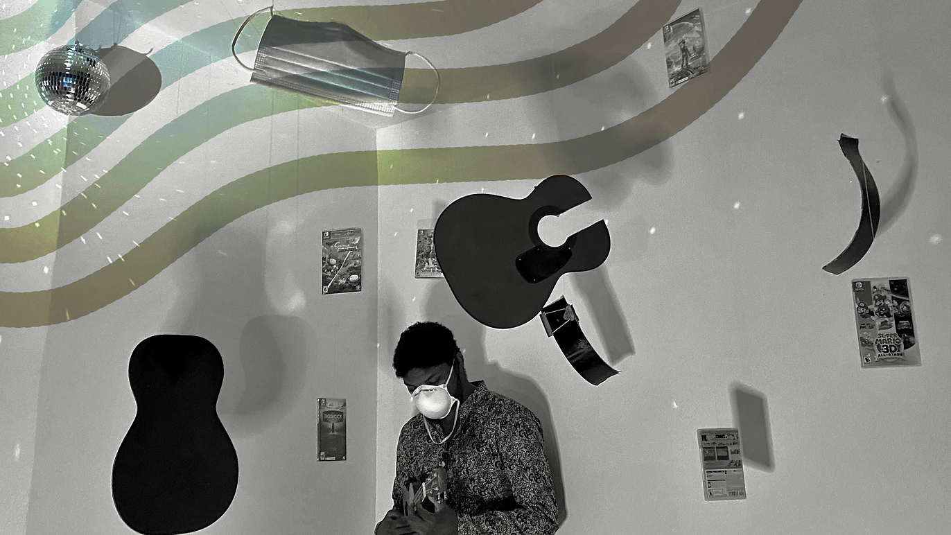 A person wearing glasses and a face mask is looking at their phone in a room with musical instruments and posters on the wall. There are hanging guitars, a disco ball, and a mask on the ceiling.