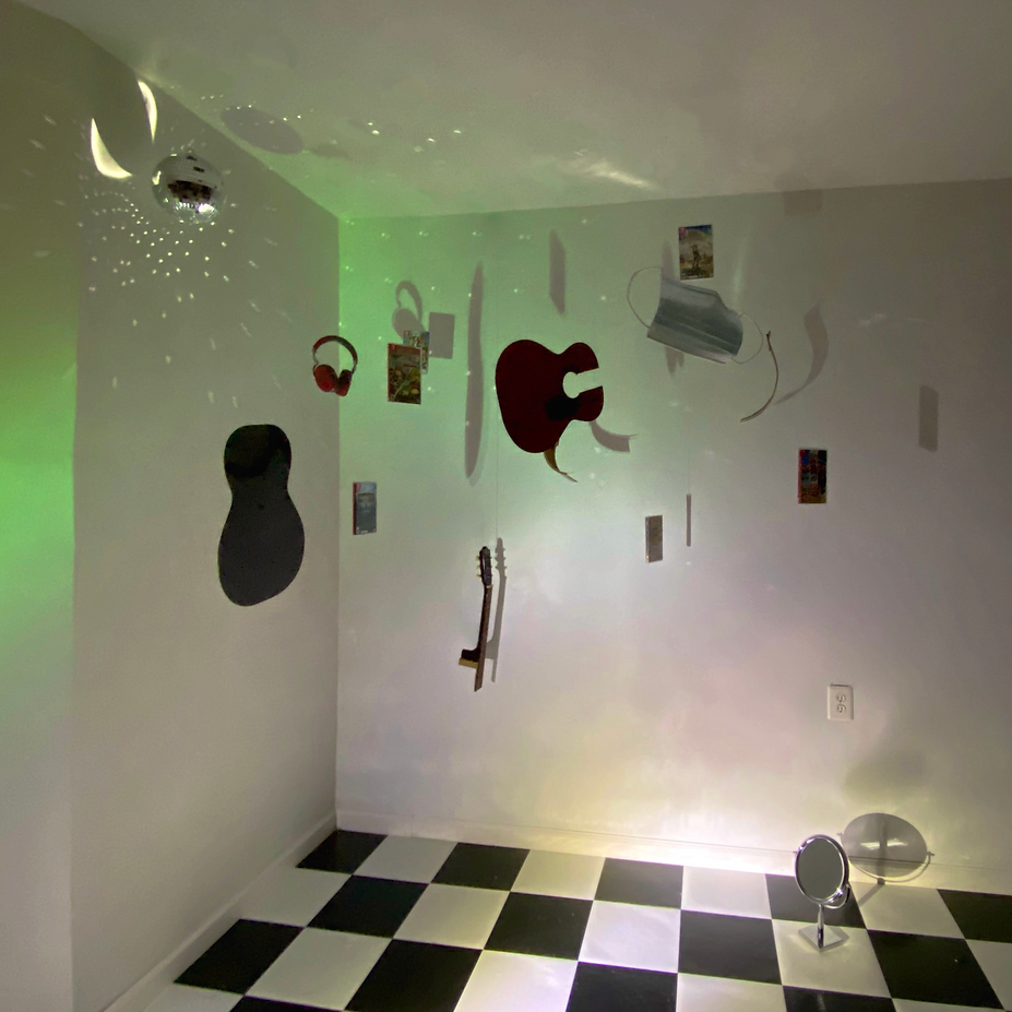 A corner of a room with a black and white checkered floor. The white wall has various objects hanging, including a large red shape, a guitar, a white mug, a pair of red headphones, and small pictures or cards. There is a disco ball hanging from the ceiling, casting small light spots on the wall and ceiling. A small round mirror on a stand is on the floor, reflecting light.