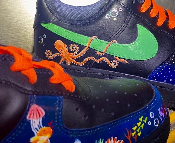 Close-up of two Nike sneakers with ocean-themed artwork. The top shoe features an orange octopus and a green Nike swoosh, while the bottom shoe depicts underwater scenes with corals, fish, and a jellyfish.