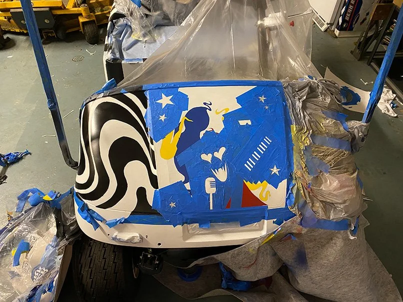 A golf cart is being spray painted with a colorful design featuring a silhouette of a woman, stars, hearts, and abstract patterns in blue, black, white, yellow, and red. The scene appears to be in a workshop with painting supplies and protective coverings.