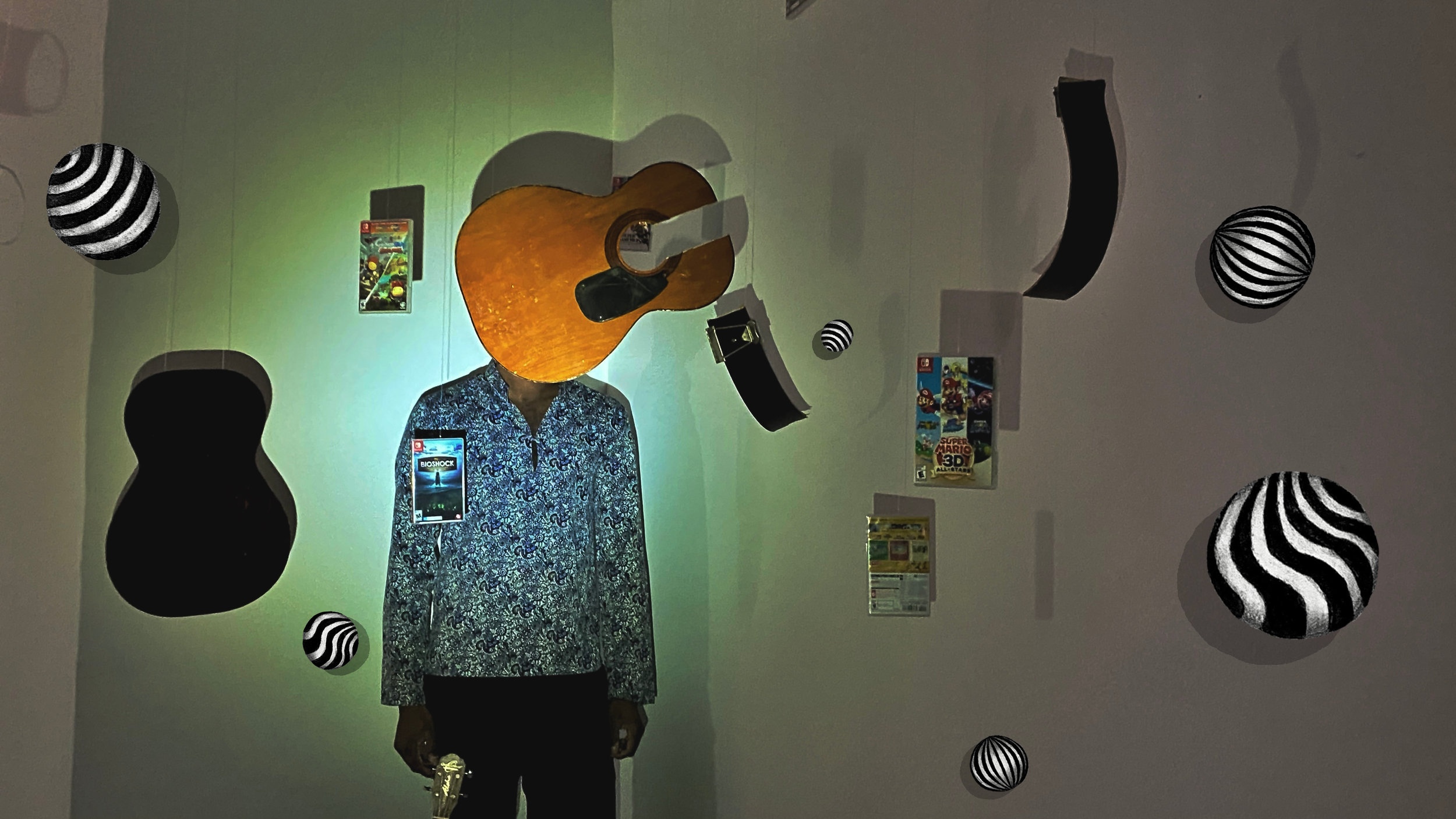 Display of a mannequin dressed in a patterned shirt, with a guitar above, surrounded by framed game cases and suspended black and white striped spheres.