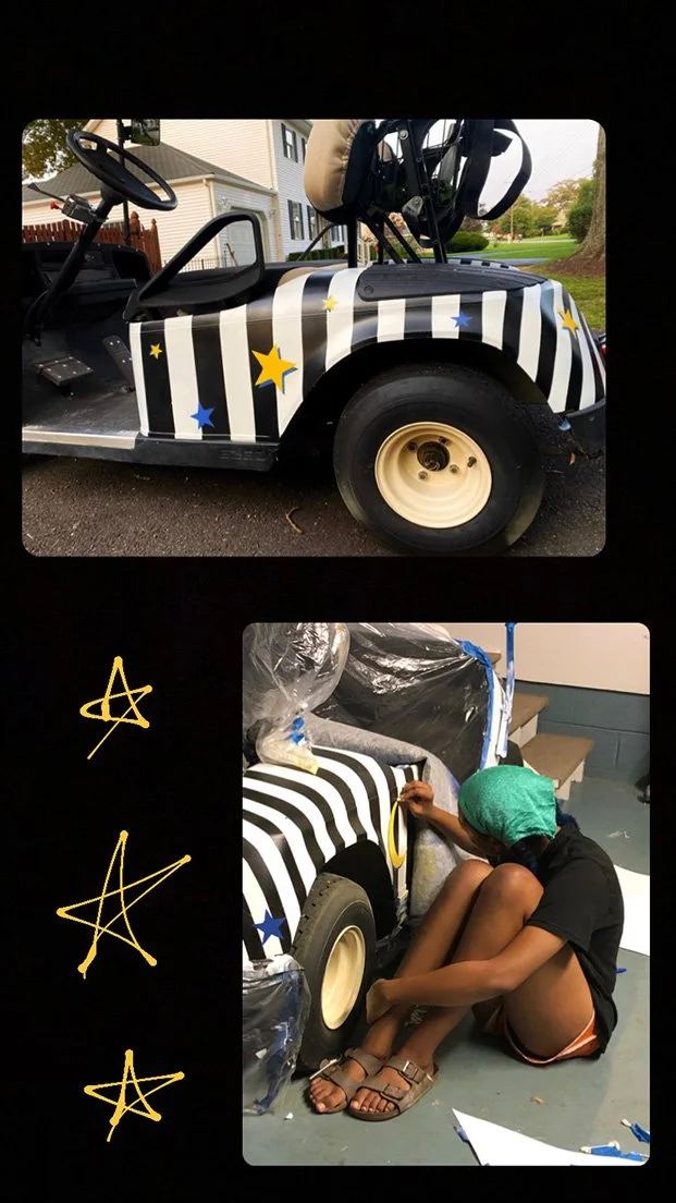 A person painting a golf cart with black and white stripes and yellow and blue star decals, sitting on the floor with a green head covering, in a garage or workshop.