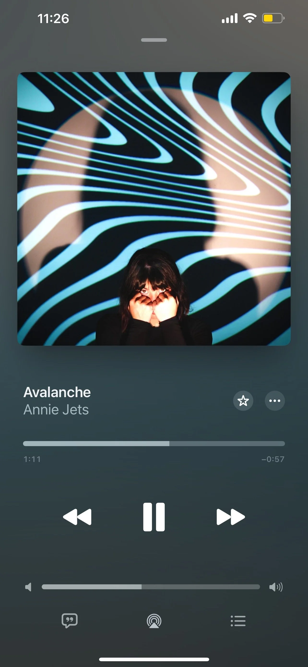 A person with dark hair covers part of their face with their hands and looks at the camera. Behind them, a wall features a large, abstract, black and light blue wavy pattern. The person is on a music app screen playing the song 'Avalanche' by Annie Jets.