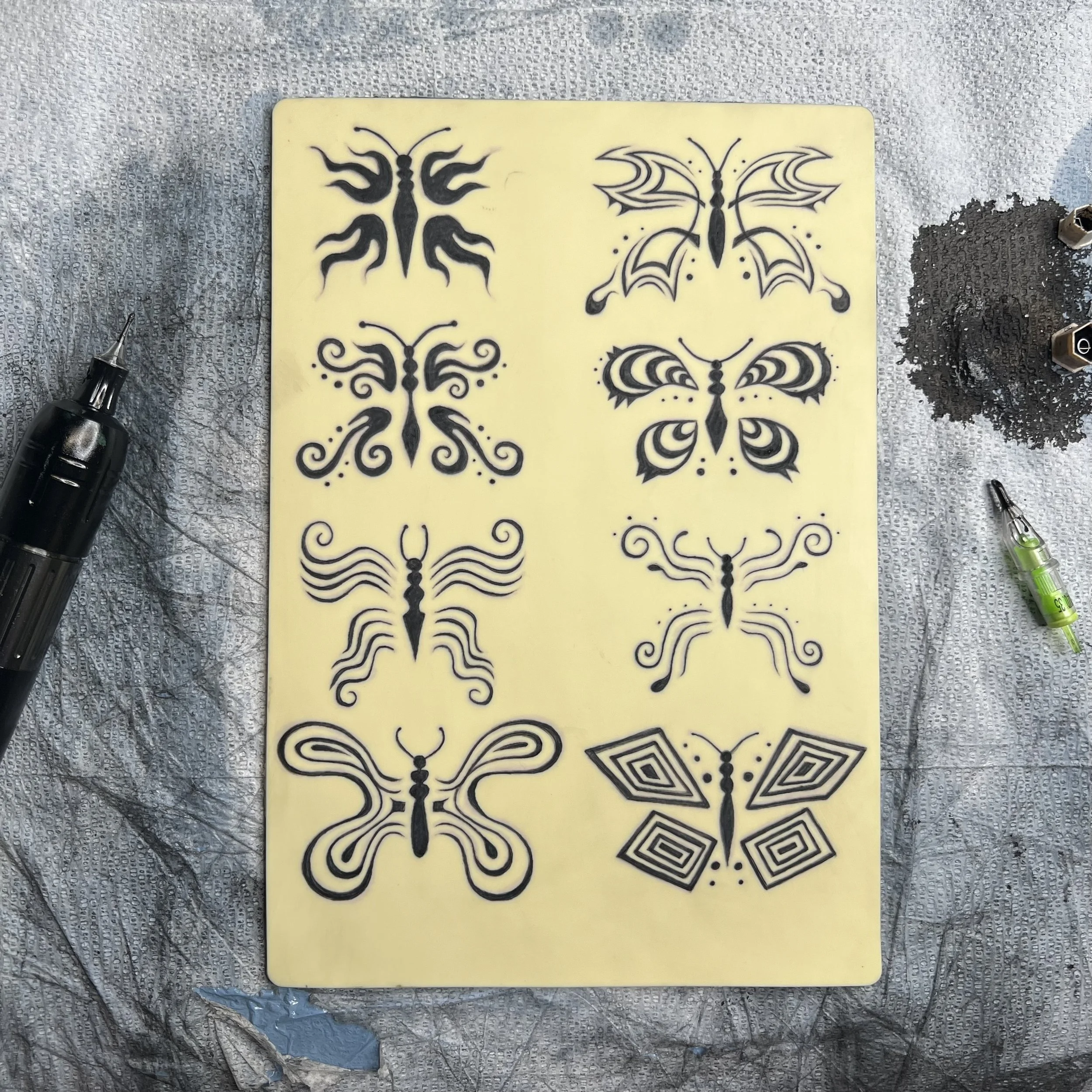 Sheet with various black butterfly tattoo designs on a yellow background, surrounded by tattoo tools and ink on a textured surface.