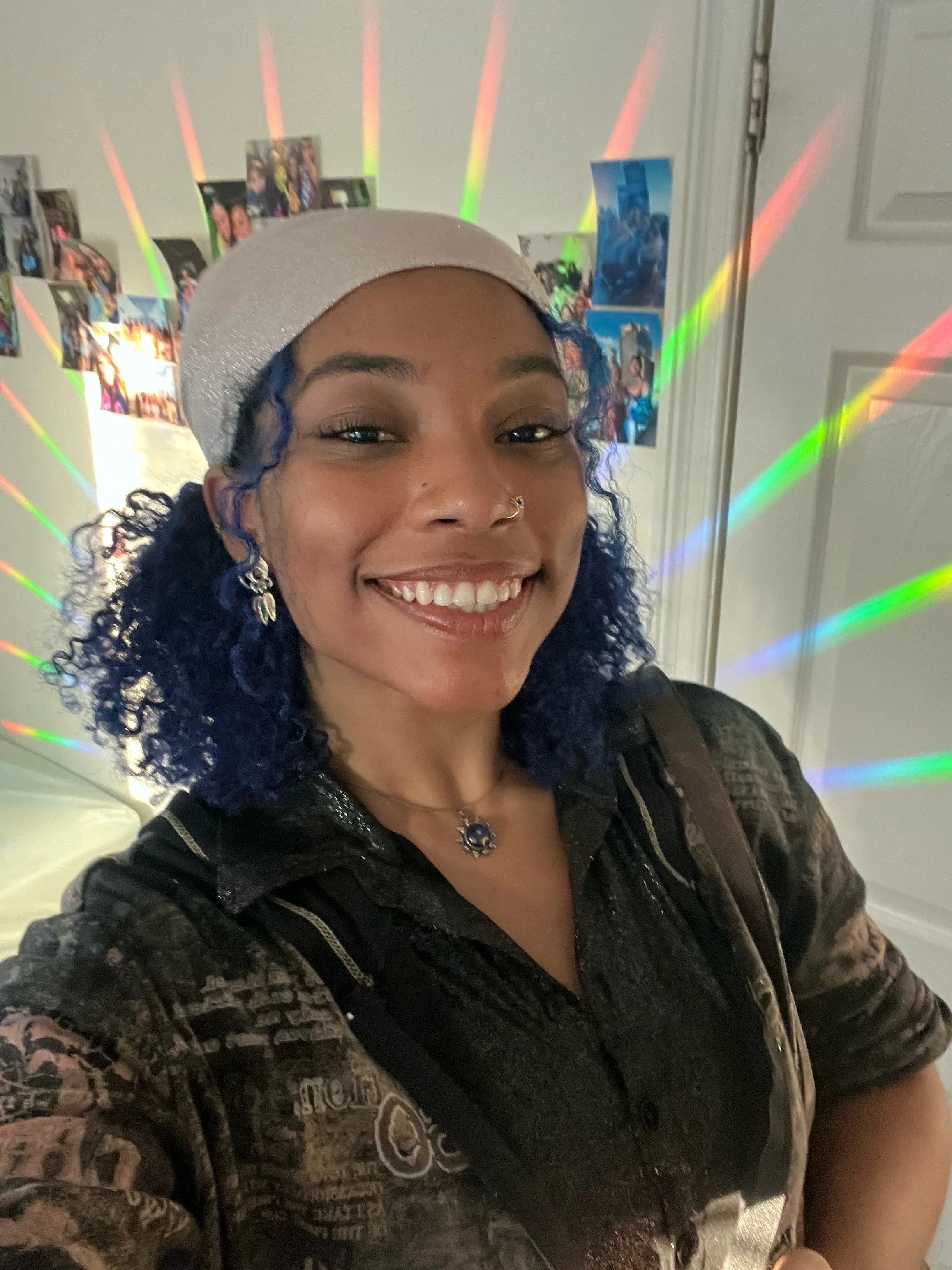 A woman with curly blue hair, wearing a beige beret, jewelry, and a black patterned jacket, smiling for a selfie in front of a wall decorated with photos and colorful light projections.