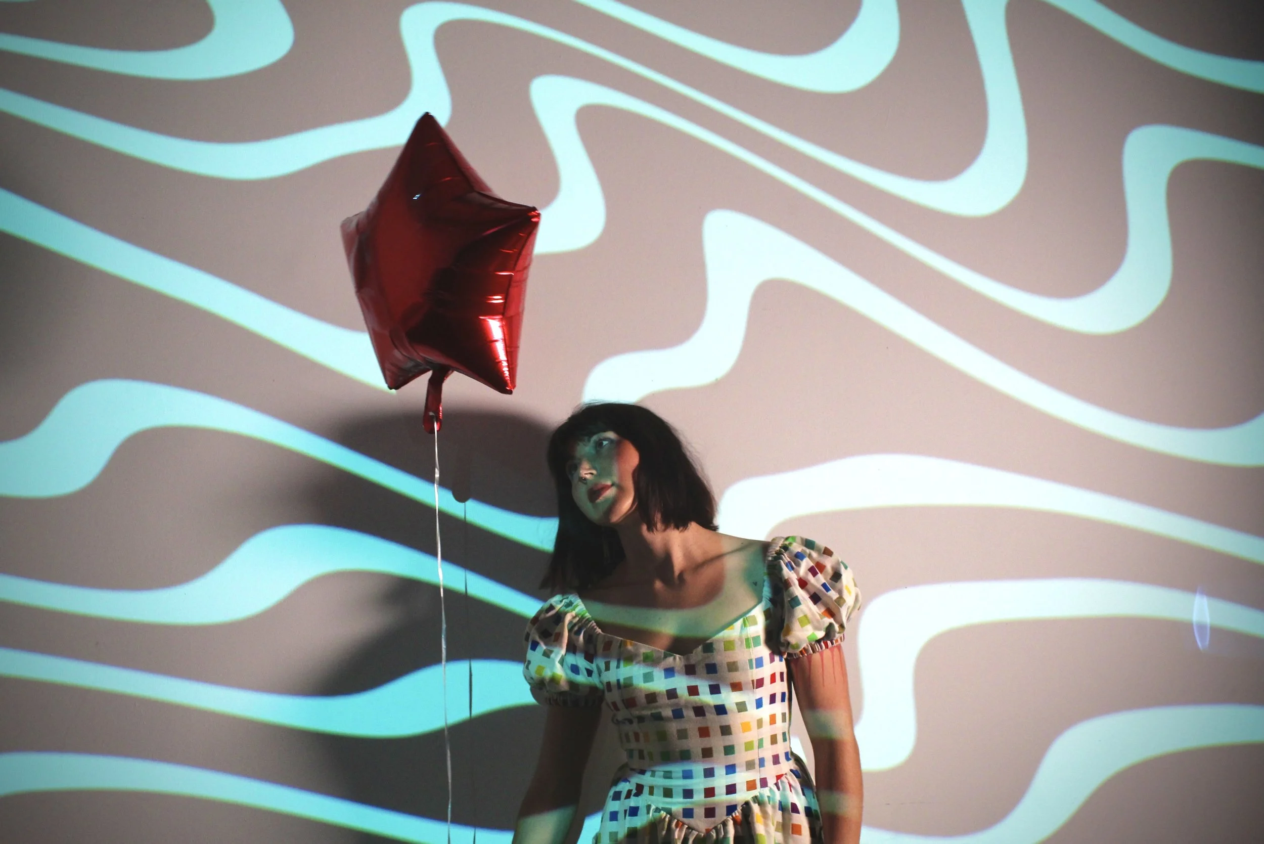A woman with shoulder-length dark hair wearing a colorful checkered dress stands in front of a background with wavy, glowing lines projected onto a wall. She holds a red star-shaped balloon.