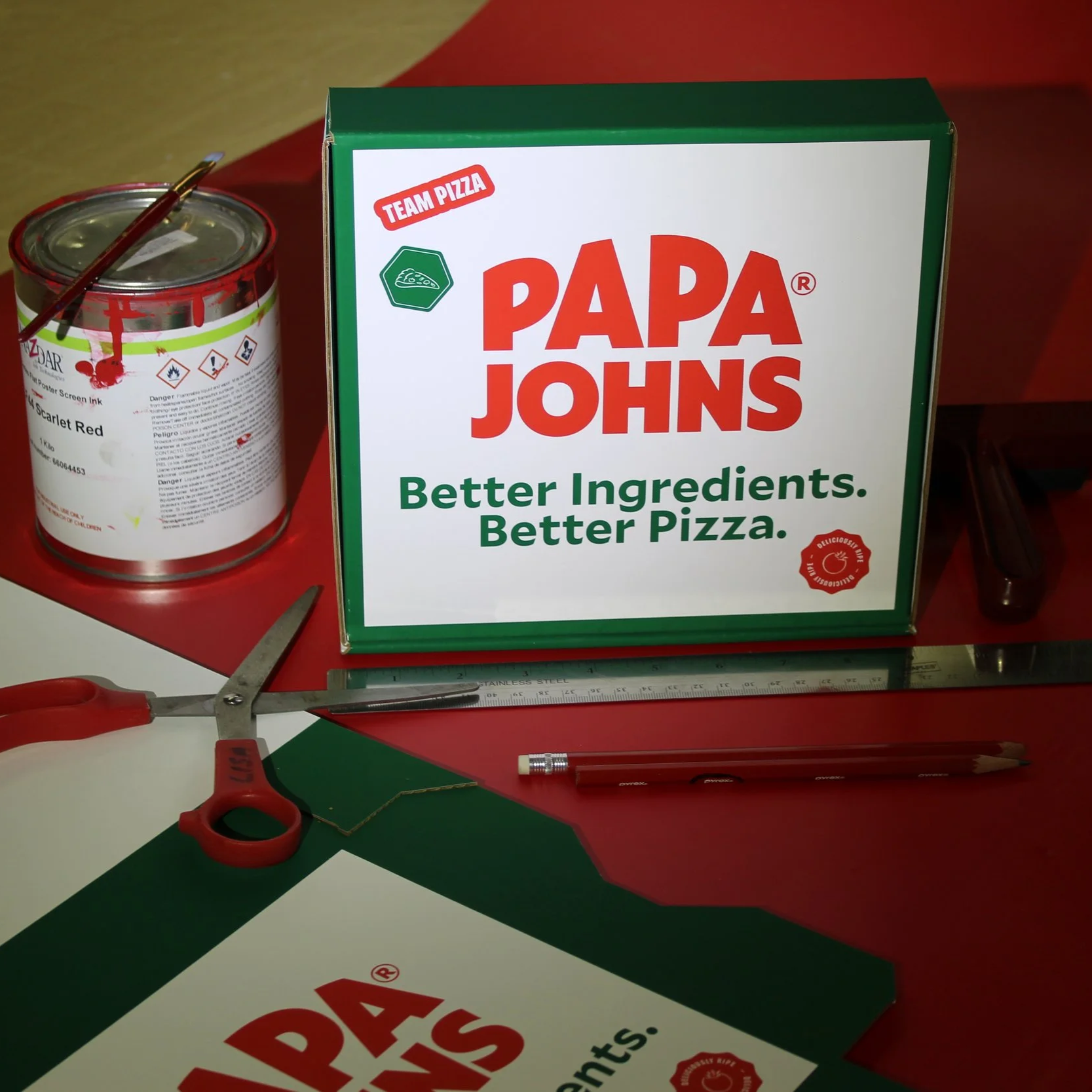 A pizza box with 'Papa Johns' branding, a pair of scissors, a red pen, a ruler, a sheet of paper, a can of red poster screen ink, a pencil, and a piece of green paper on a red surface.