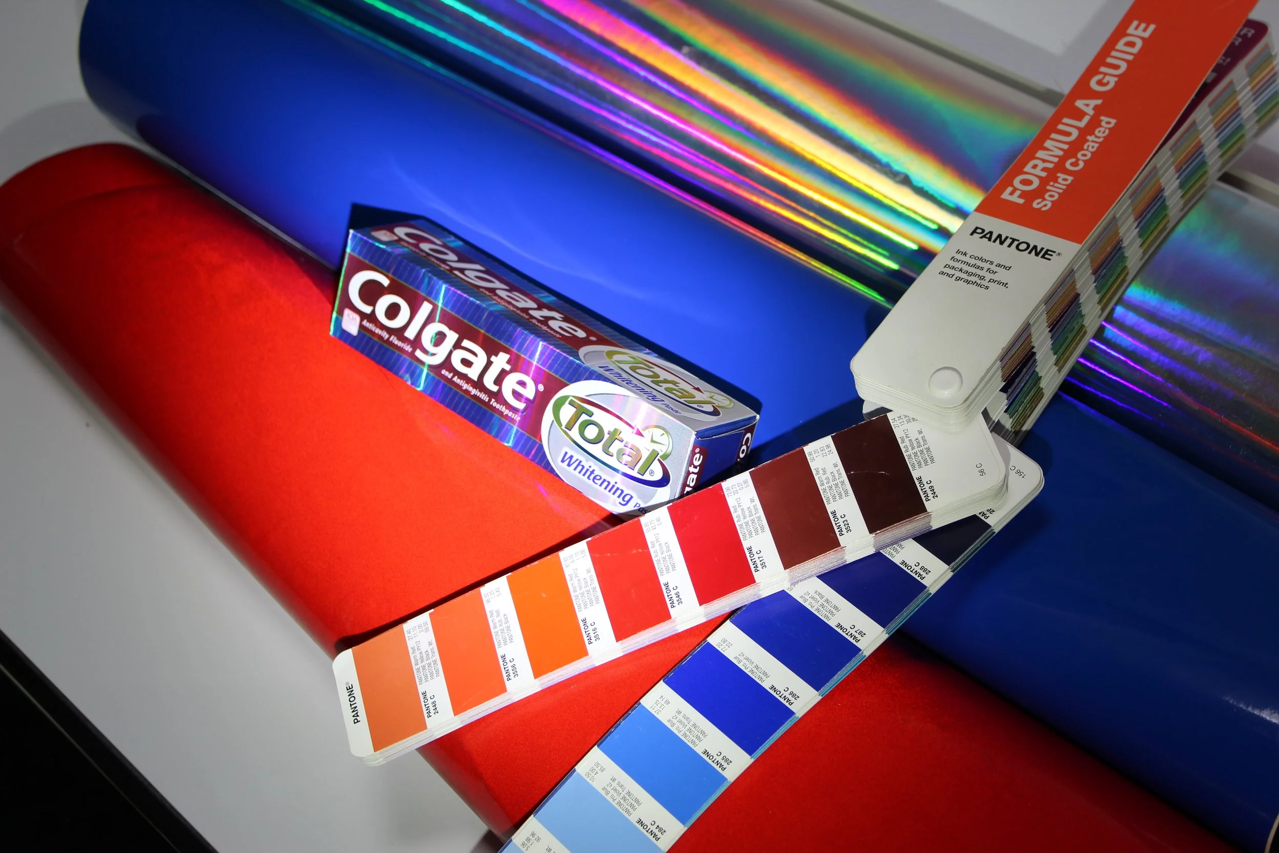 Colorful holographic laminated paper sheets, an orange paint guide strip, and a Pantone color swatch book on a white surface.