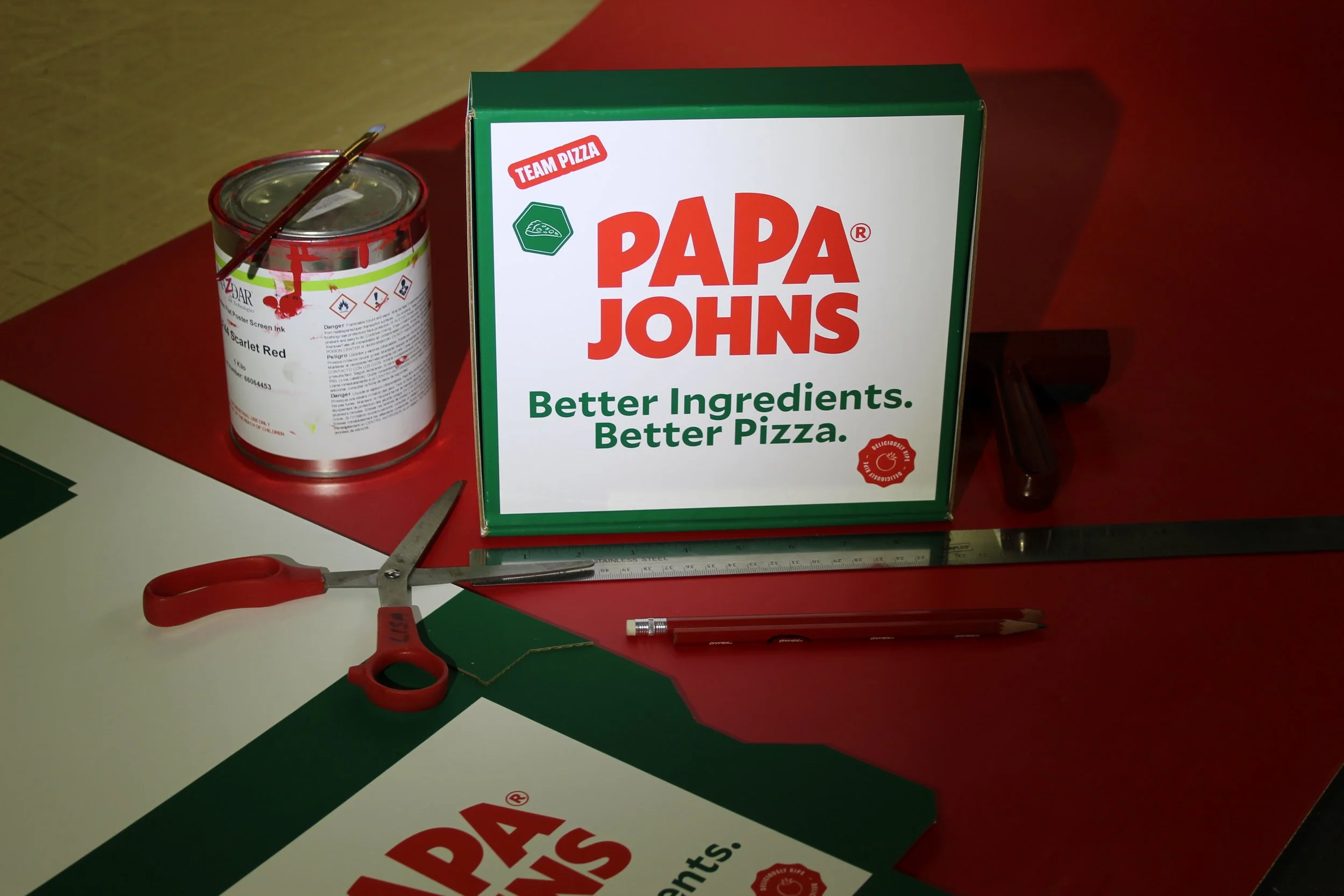 A cardboard pizza box with the Papa John's logo and slogan, a can of red screen ink with warning symbols, a pair of scissors, a utility knife, a ruler, and a pen on a table.