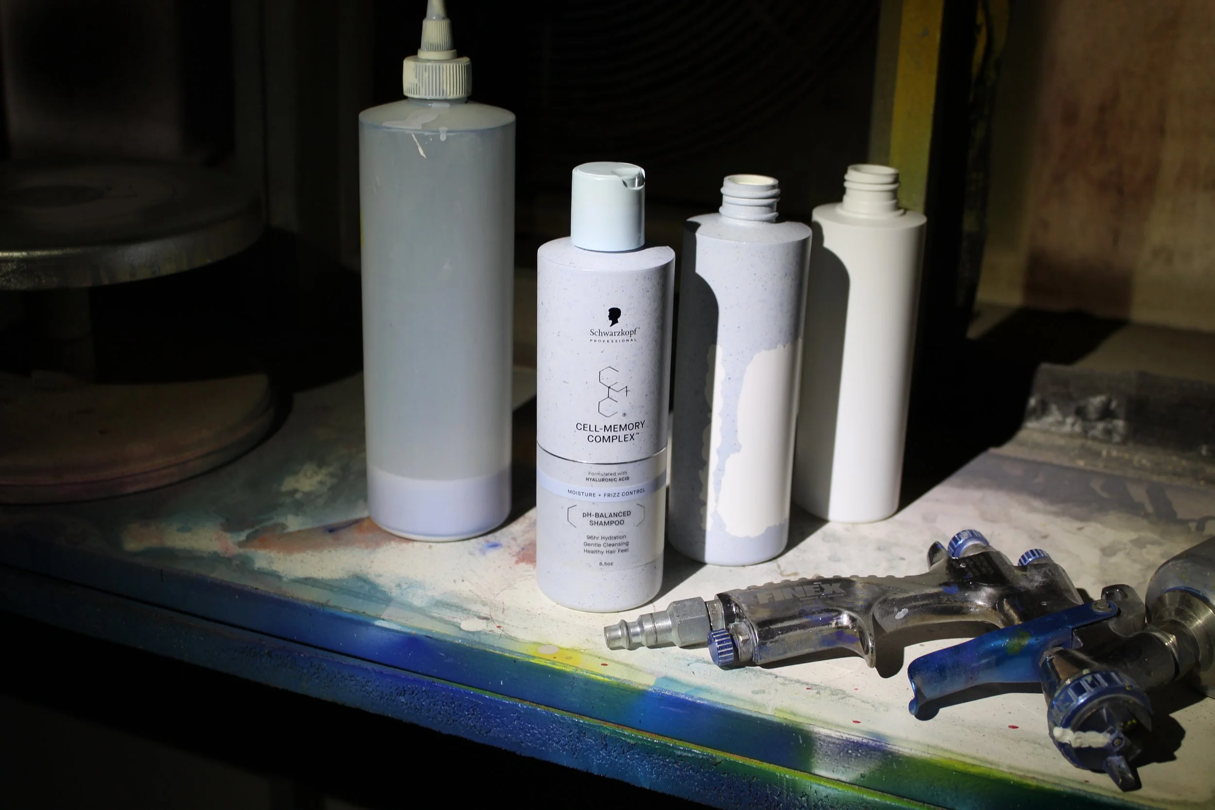 A row of white bottles, including Schwarzkopf Cell-Memory Complex shampoo, are arranged on a painted shelf with spilled paint and spray nozzles around them.