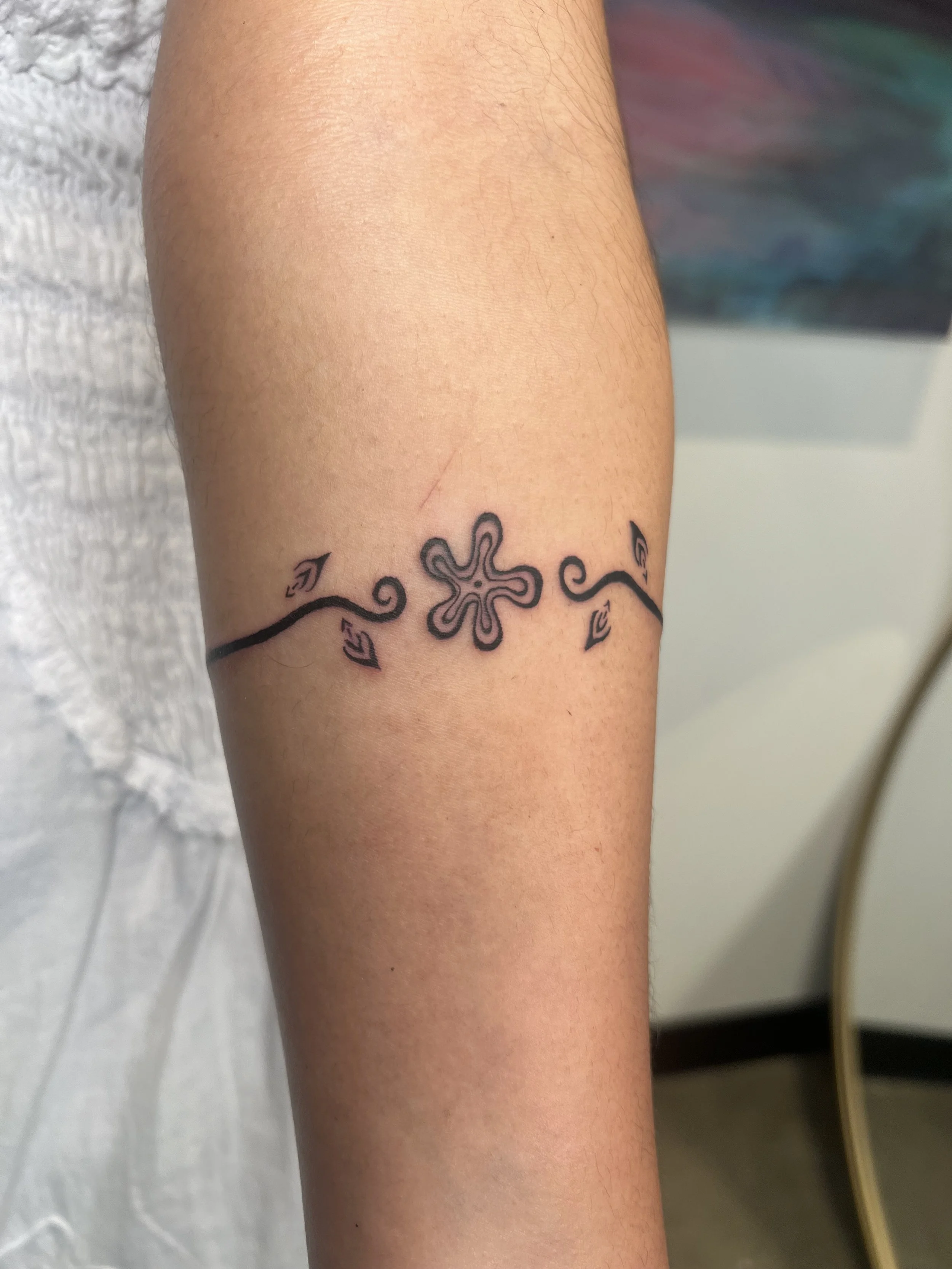Close-up of a person's forearm with a black tattoo of a symmetrical abstract flower with swirling lines and leaf-like shapes.