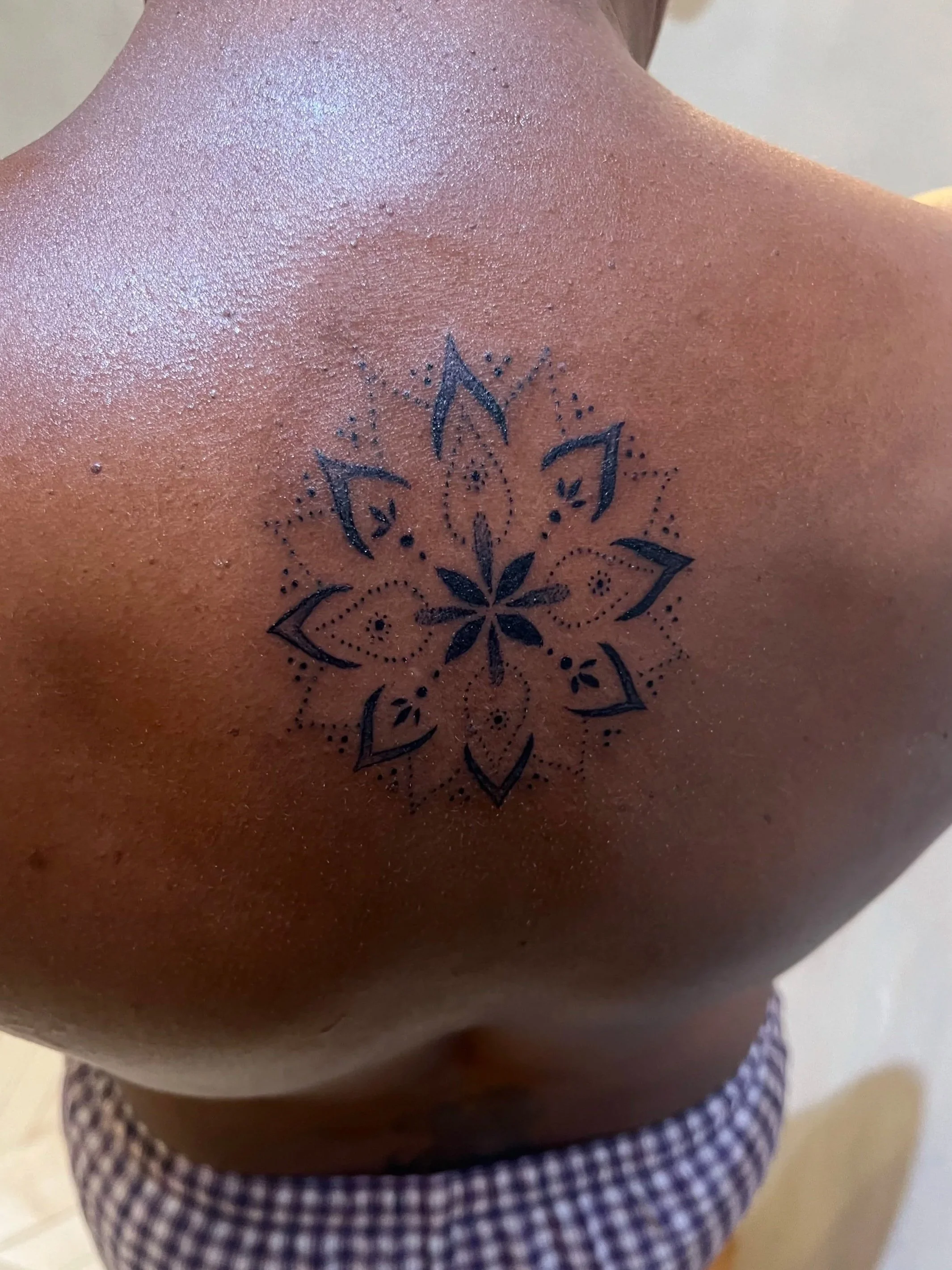Close-up of a person's upper back and shoulder showing a black arm tattoo with a geometric floral pattern.