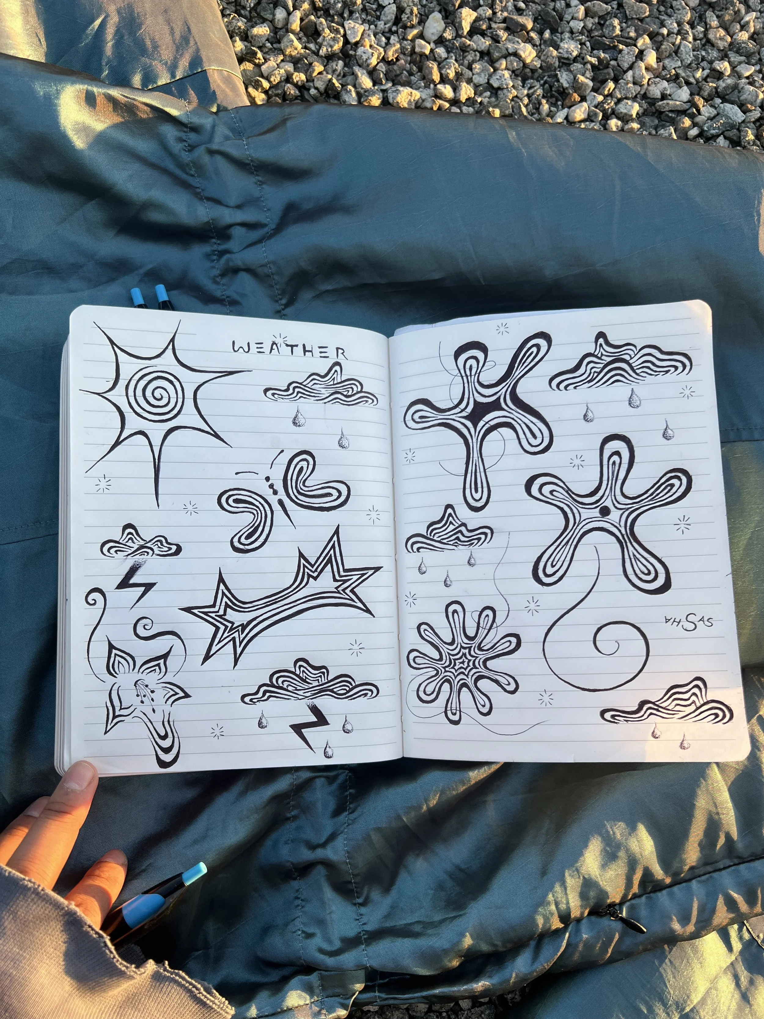 Open notebook on the person's lap with black line drawings of weather symbols, including a sun, clouds, raindrops, snowflakes, and lightning.