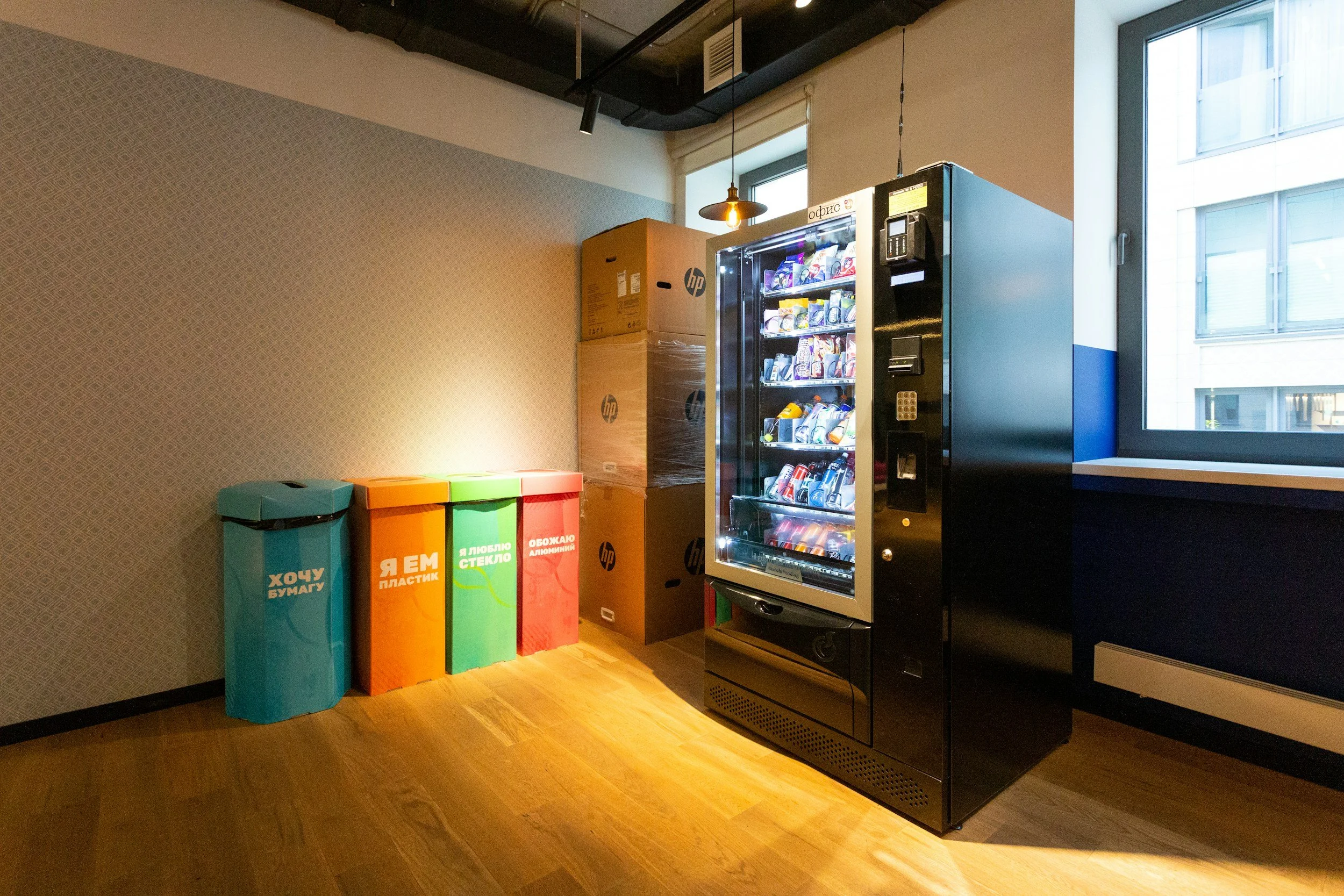 Interior of an office or break room with a vending machine, colorful recycling bins, and stacked cardboard boxes near the wall.