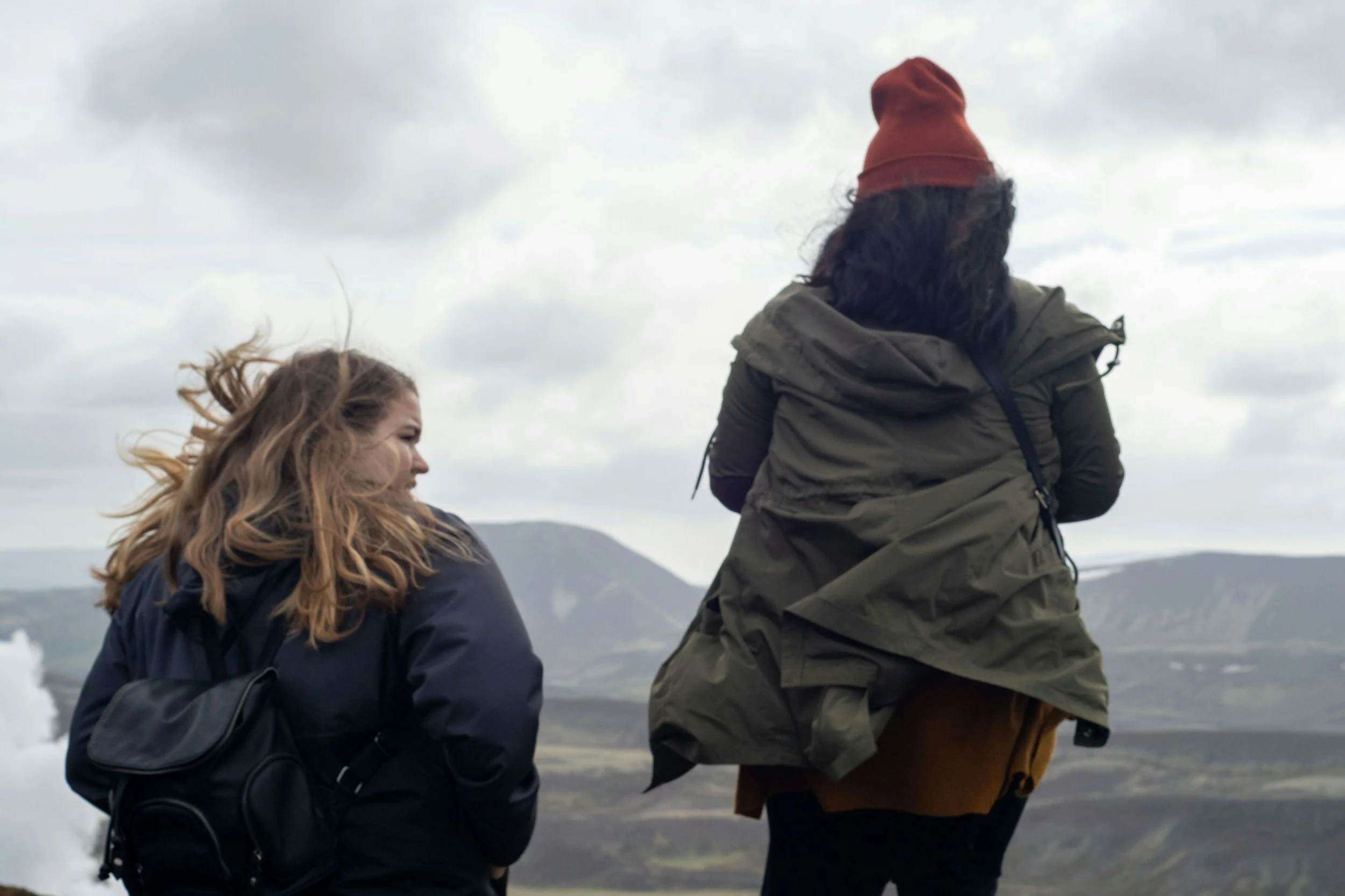 Two women with long hair and jackets are outdoors near a body of water with mountains in the background, under cloudy skies.