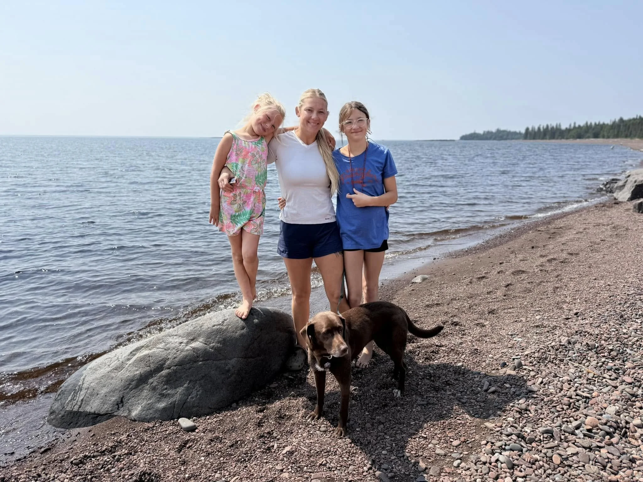 Three young girls and a dog stand on a rocky beach near the water with a forested area in the background under a clear sky.