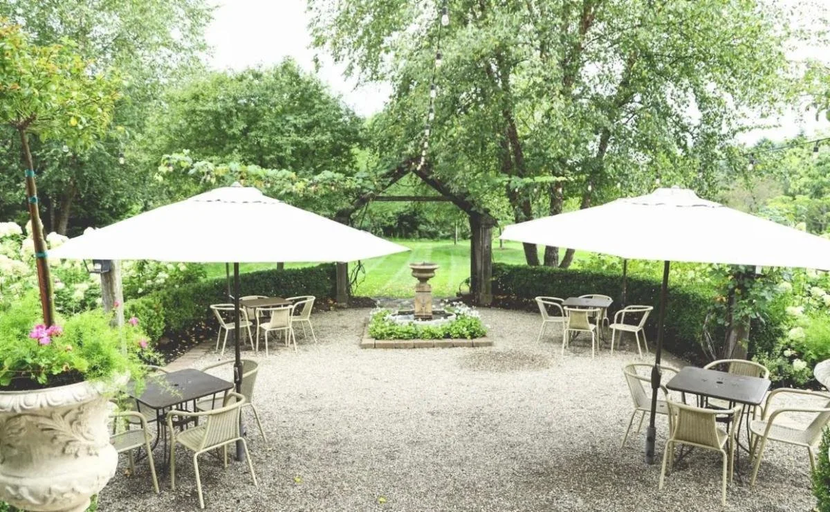 Outdoor patio with gravel ground, two large white patio umbrellas, multiple tables and chairs, surrounded by green trees and bushes, with a fountain in the background.