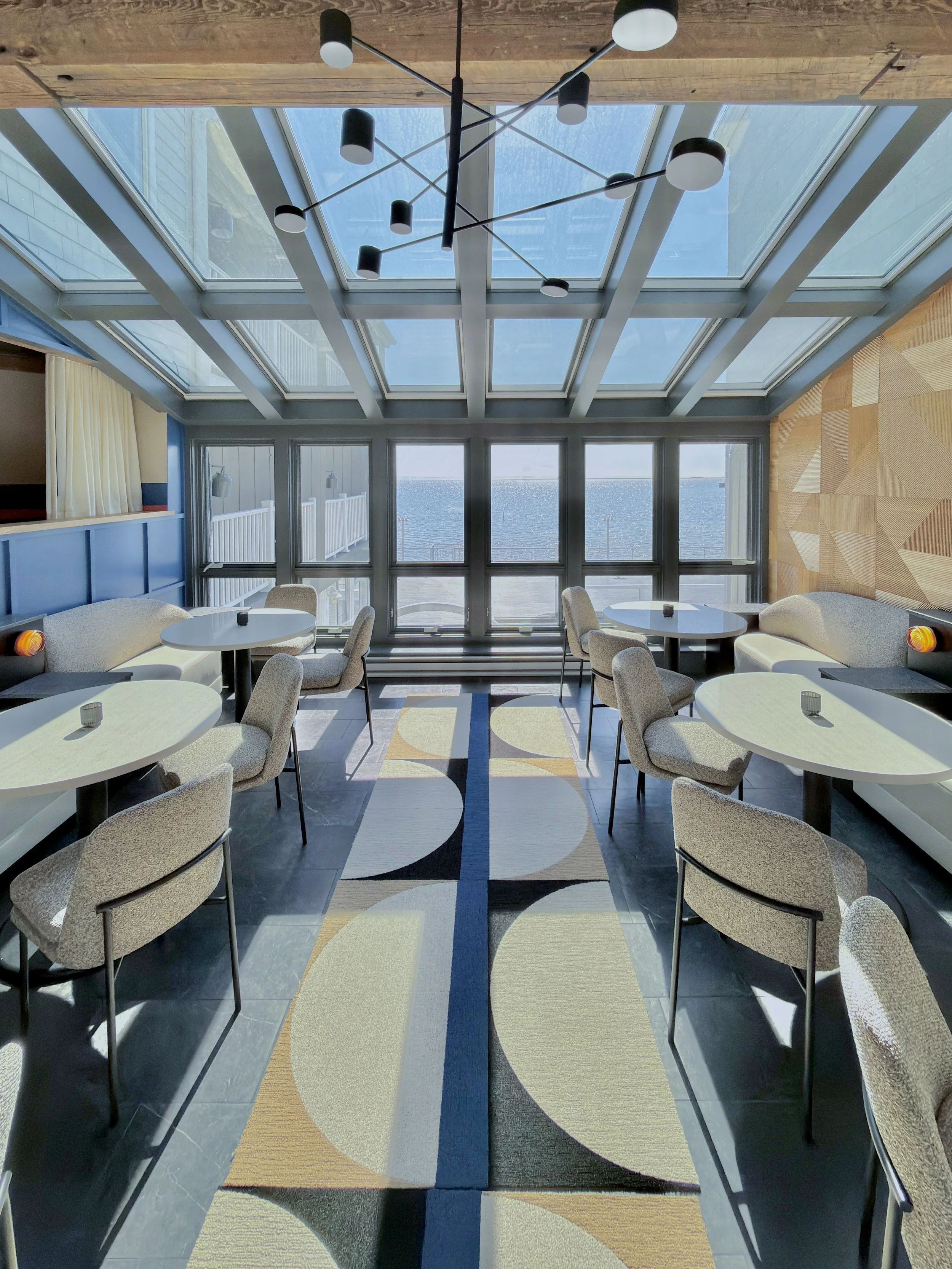 A modern indoor dining area with round tables and upholstered chairs. Large windows provide a view of the water and sky outside. A geometric patterned rug is on the floor, and a contemporary chandelier hangs from a wooden ceiling with a skylight.