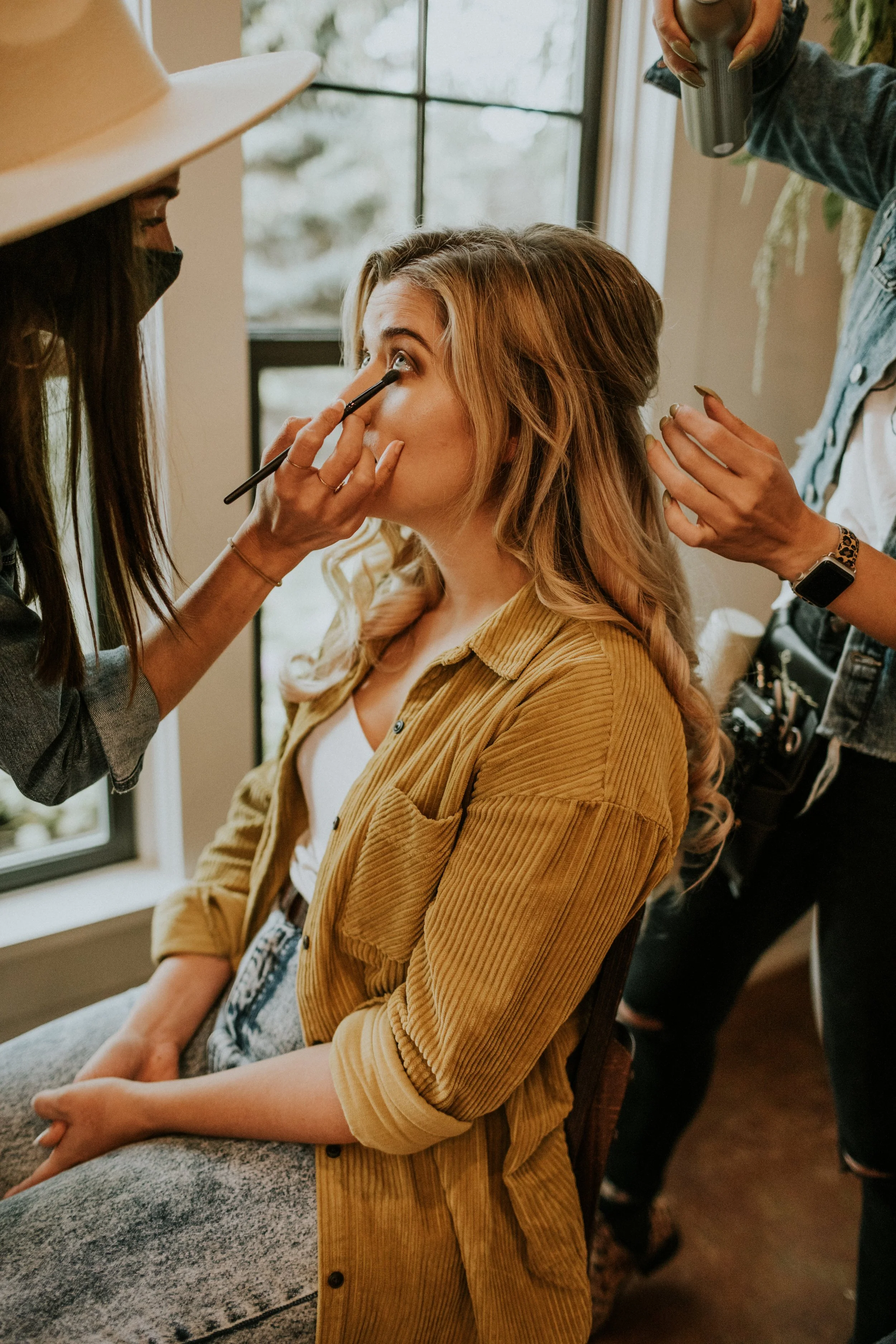 A woman is getting her makeup done by a makeup artist inside a room with natural light coming through a window. The woman is sitting with her hands resting on her lap, wearing a mustard-colored corduroy jacket, and has long, curled hair. The makeup artist, partially visible, is applying makeup around her eye with a brush.