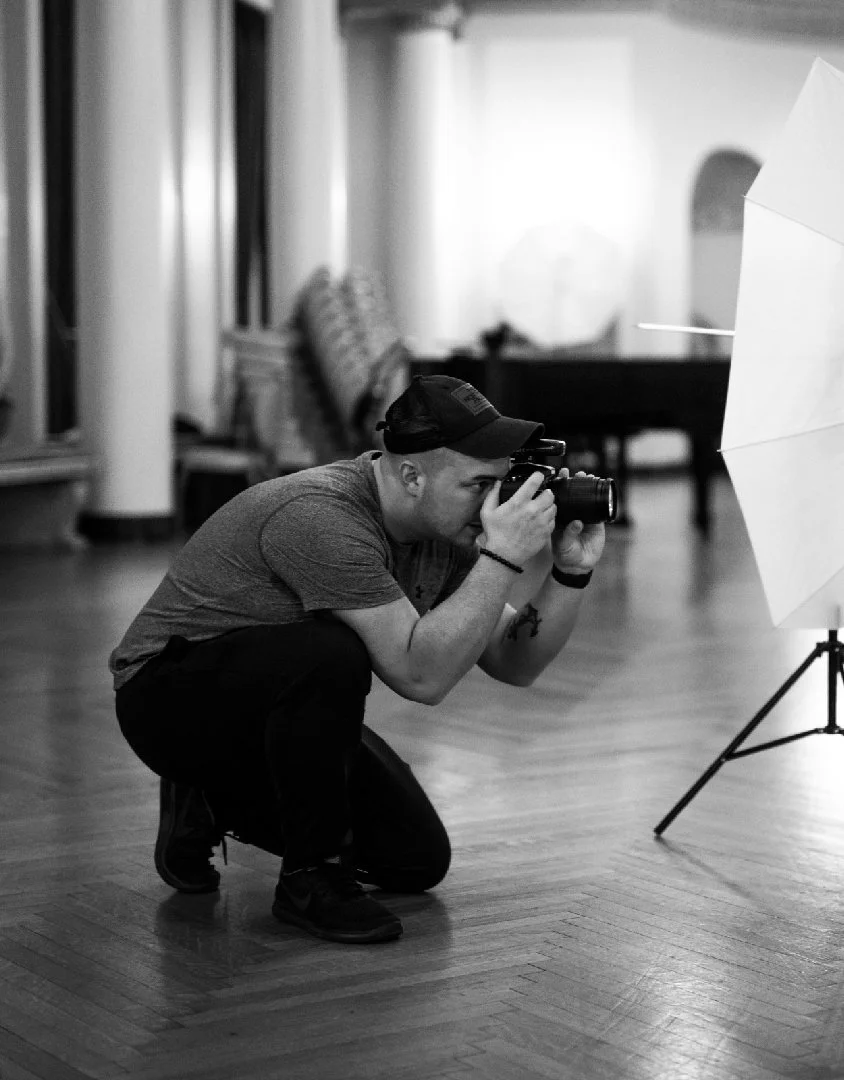 A man crouching on the floor taking a photograph with a camera, indoors with a photography umbrella nearby.