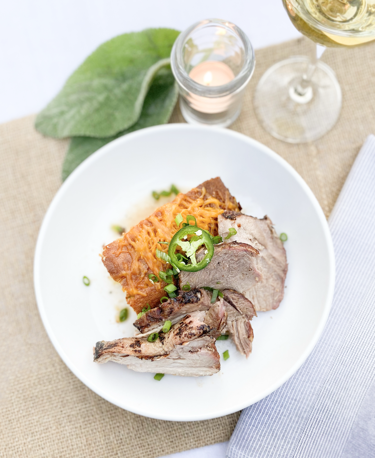 A white plate with sliced roast beef, cheesy baked potato, and green garnishes on top. In the background, a small candle in a glass holder, a large leaf, and a glass of white wine on a beige tablecloth.