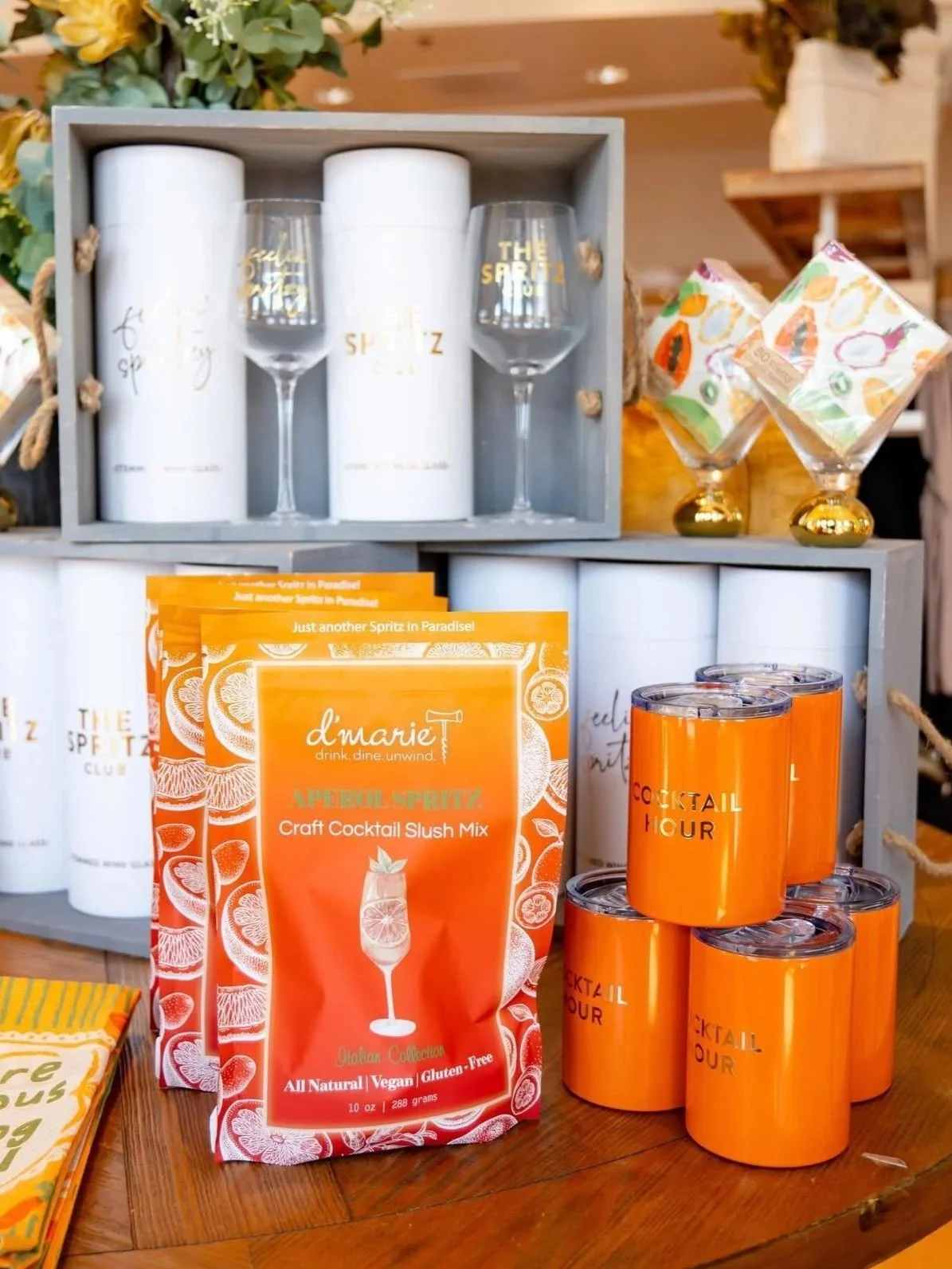 Display of craft cocktail slush mix packets, orange cocktail cups labeled 'Cocktail Hour', wine glasses, and decorative packaging with floral designs, all arranged on wooden shelves.