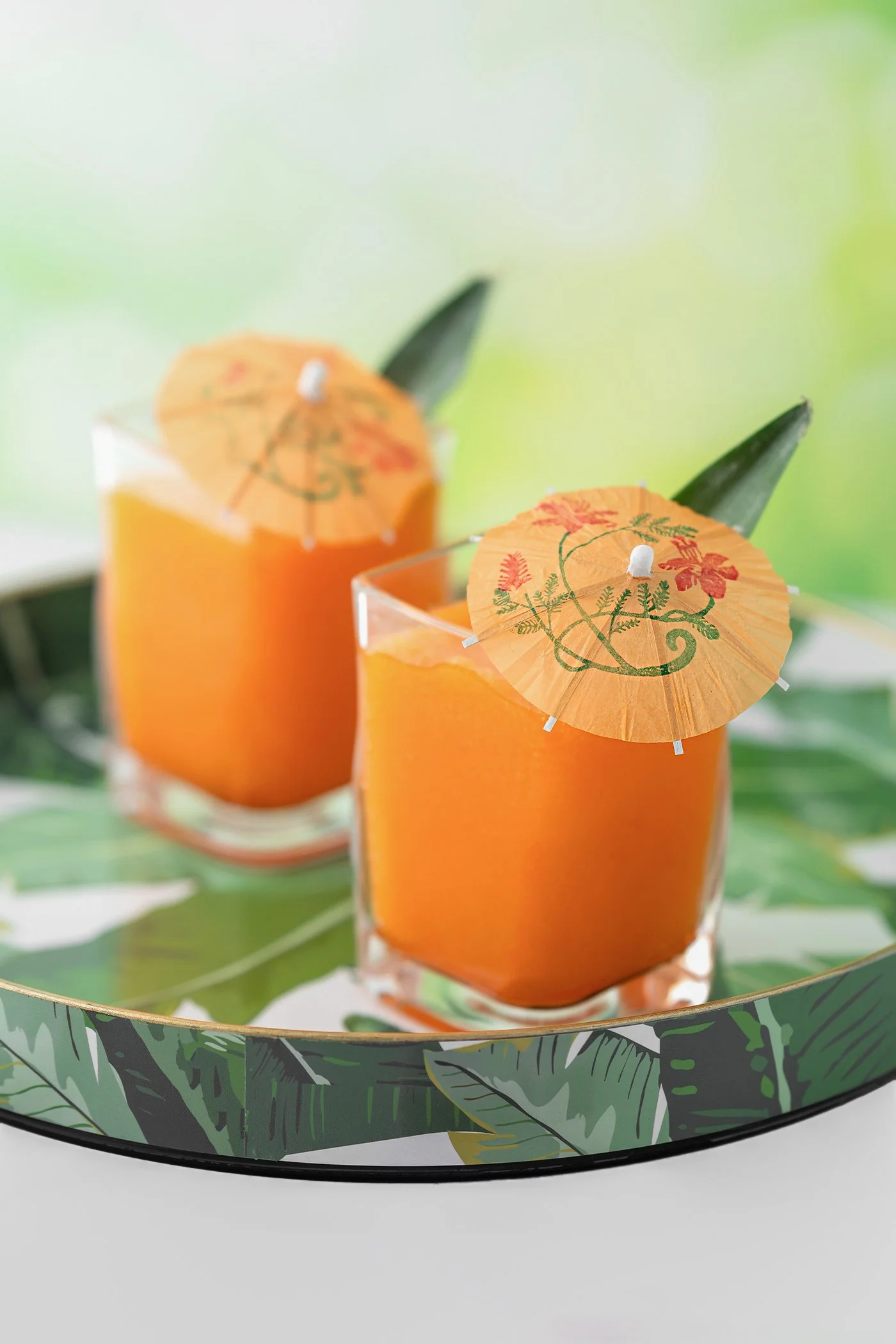 Two glasses of orange juice with paper umbrellas and green leaves in the background, on a decorative tray.