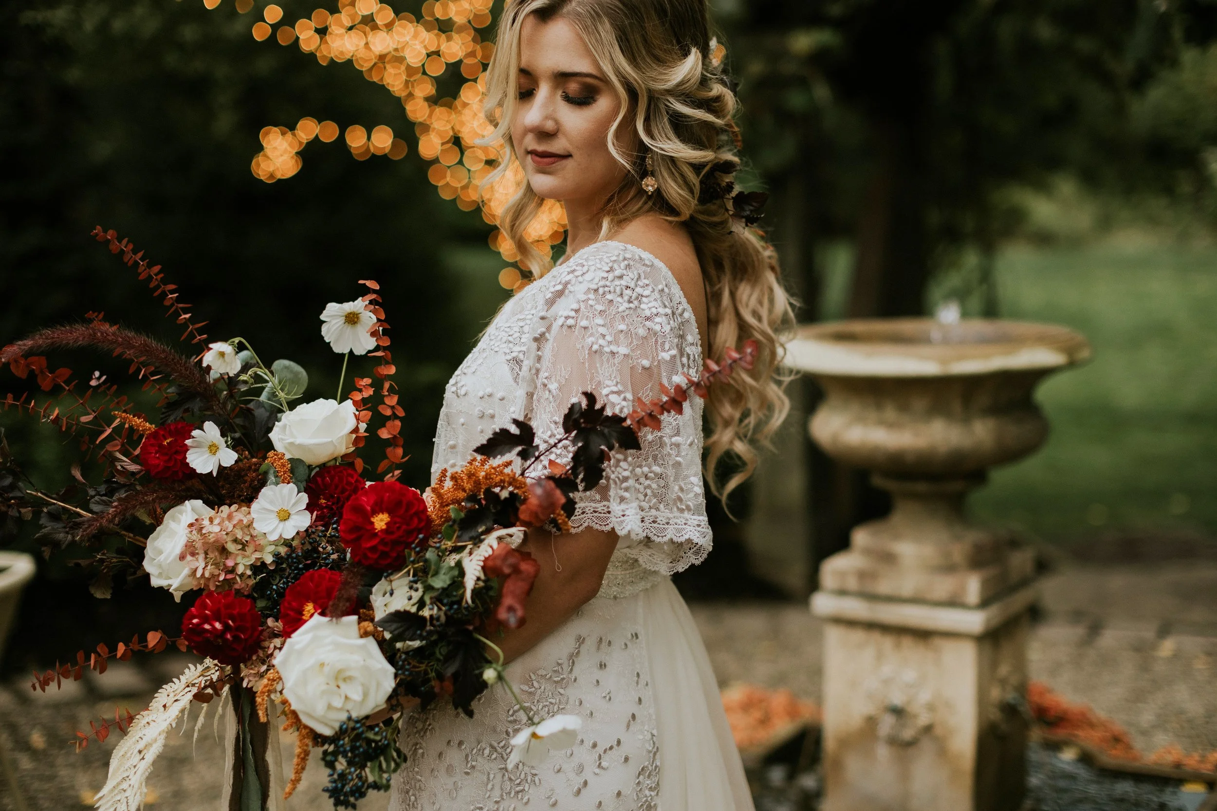 A bride with long curly blonde hair, wearing a lace wedding dress, holding a large bouquet of flowers, standing outdoors with blurred greenery and bokeh lights in the background.