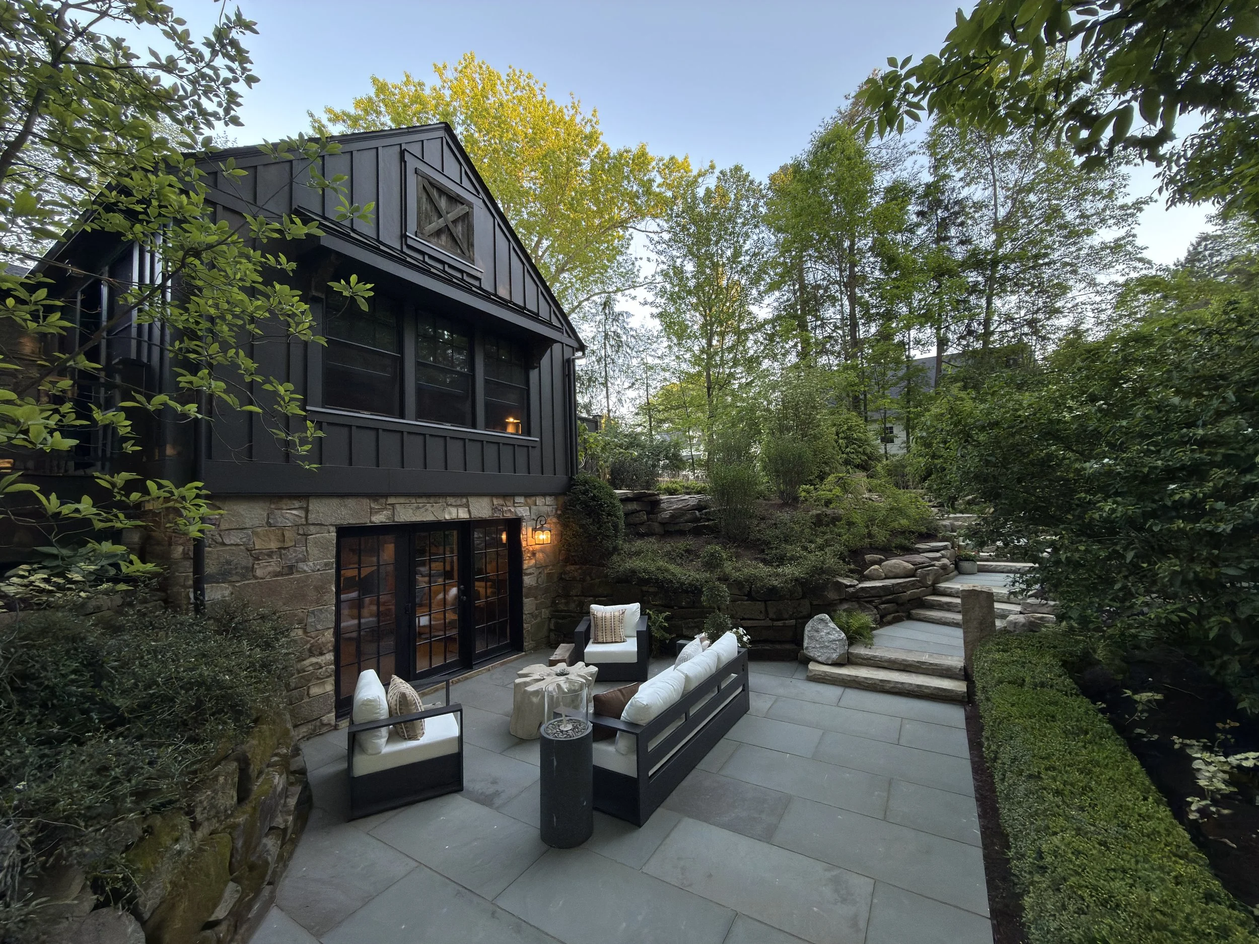 A backyard patio with outdoor furniture including a sofa and chairs with cushions, surrounded by greenery and trees, with a house in the background.
