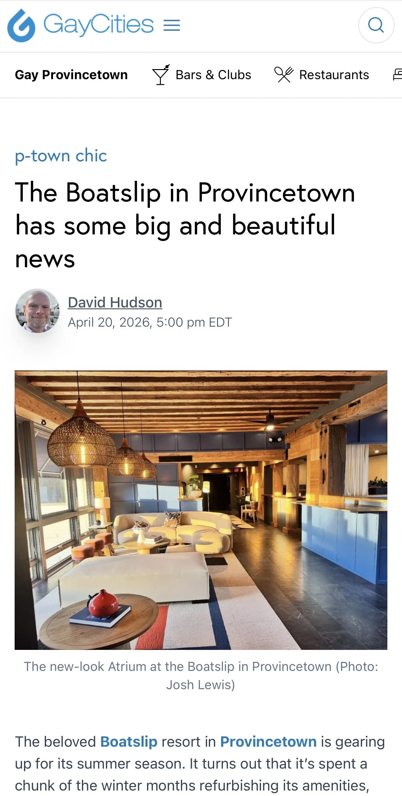 Interior view of a hotel lobby with modern decor, wooden ceiling beams, hanging pendant lights, large windows, a beige sectional sofa, colorful area rug, and a blue reception desk.