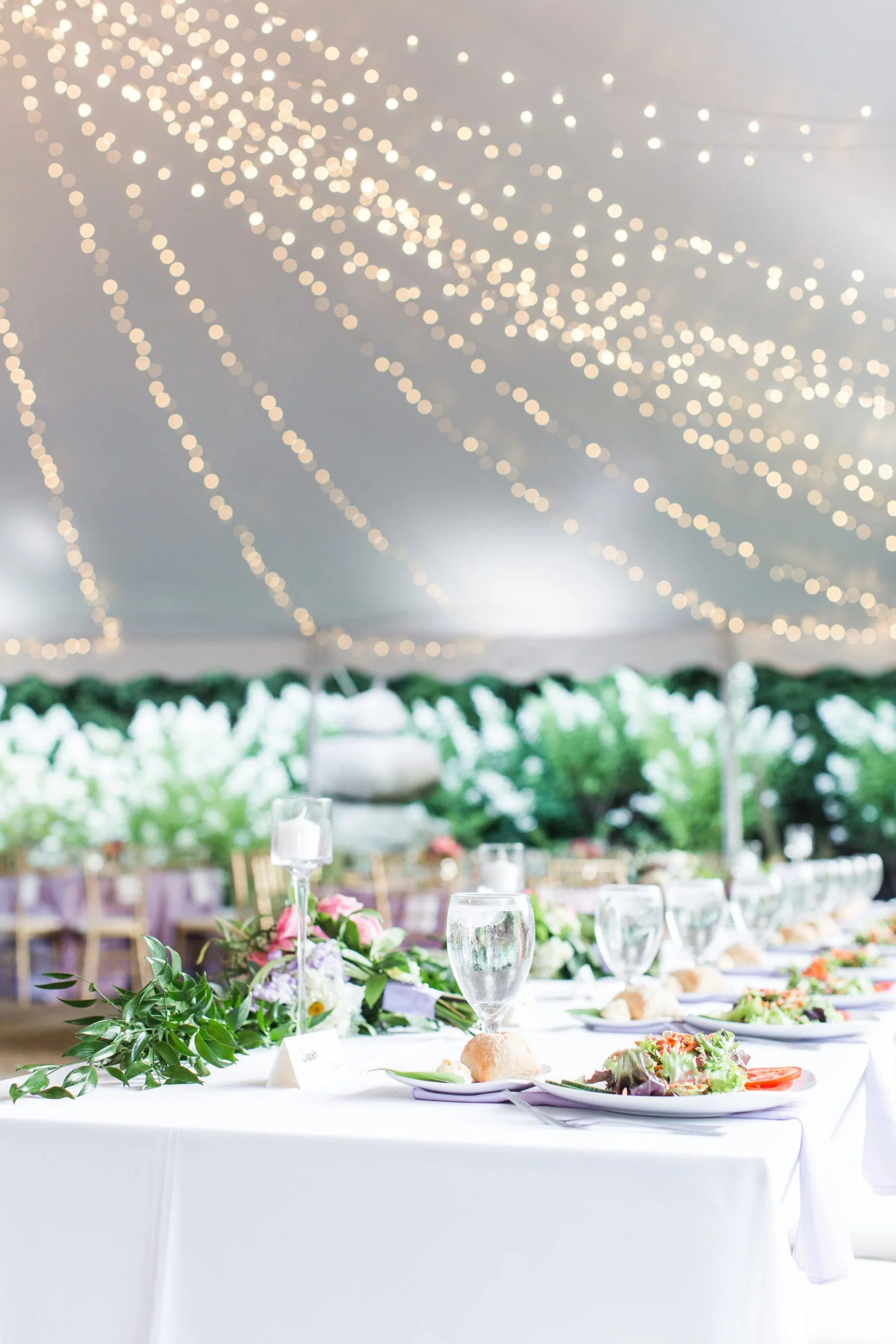 A decorated event table under a tent with floral centerpieces, glasses, plates of salad and bread, and a blurred background of trees.