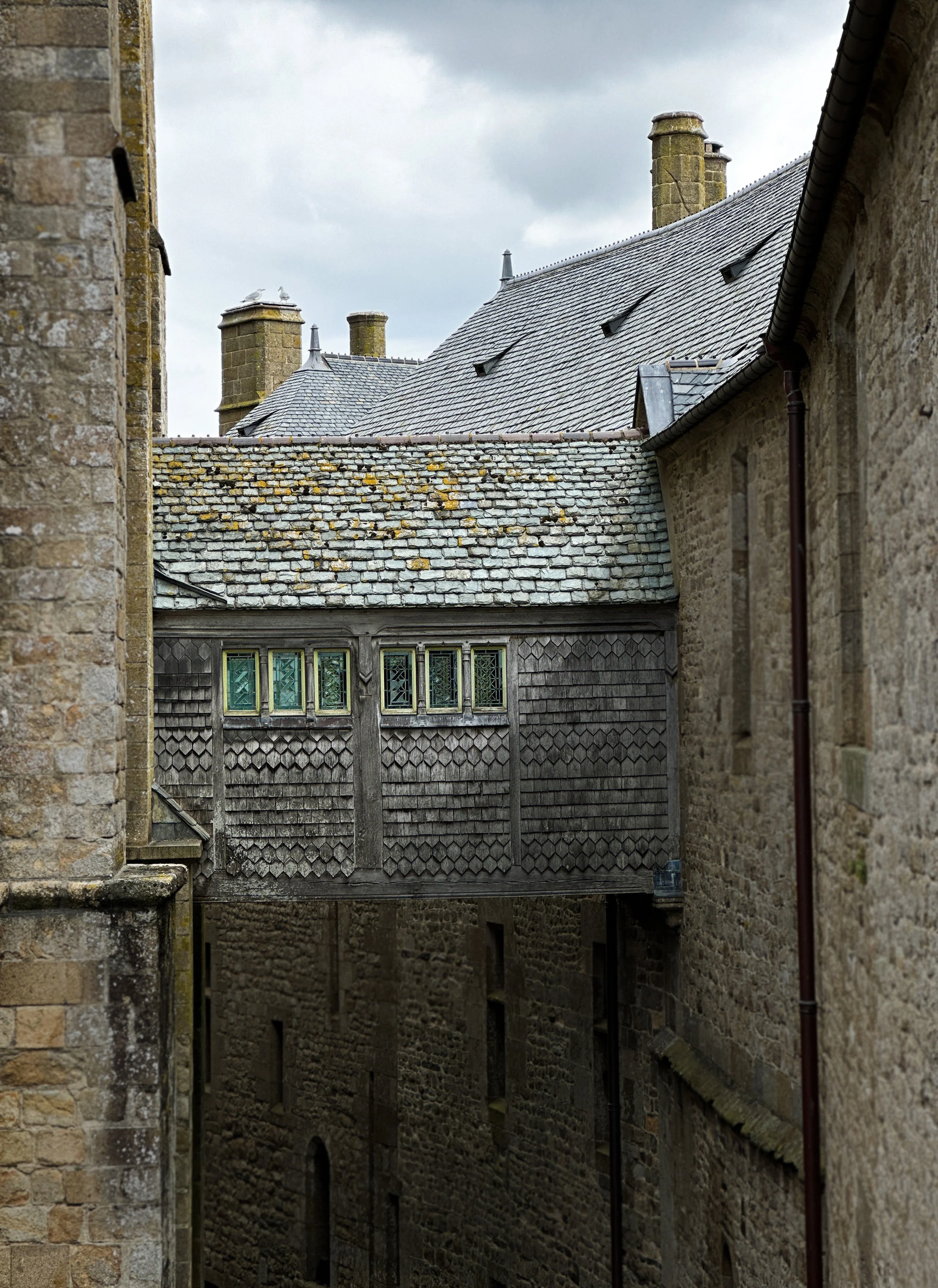 A narrow alleyway between old stone buildings with chimneys and a wooden balcony. Overcast sky.