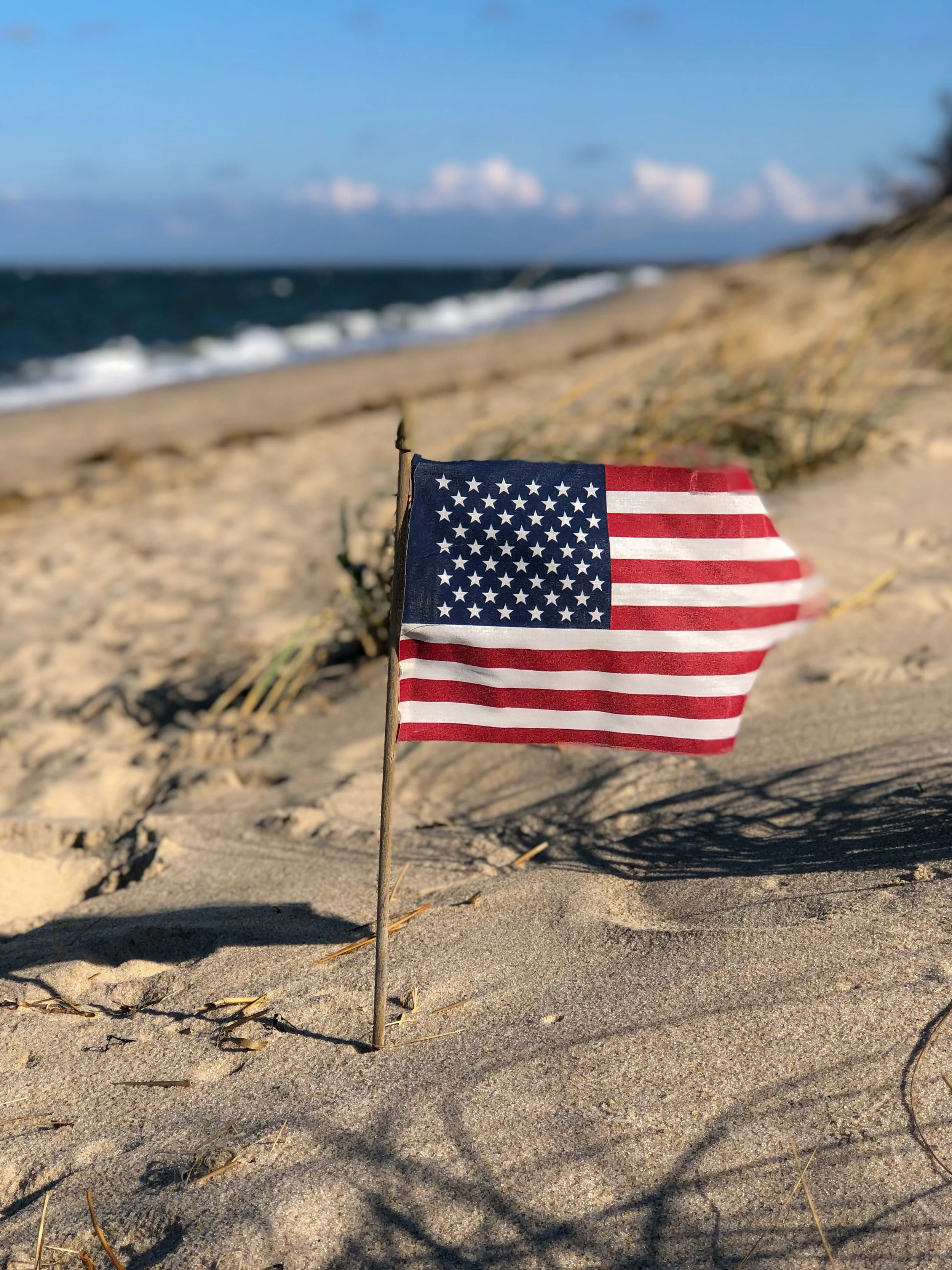 Small American flag on a stick planted in sandy beach with ocean waves and blue sky in the background.