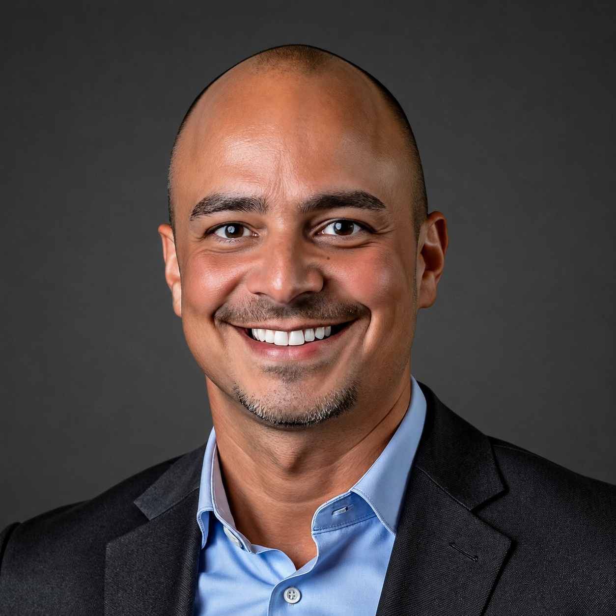 Professional headshot of a smiling man with a shaved head, facial hair, wearing a black blazer and light blue shirt, against a dark background.