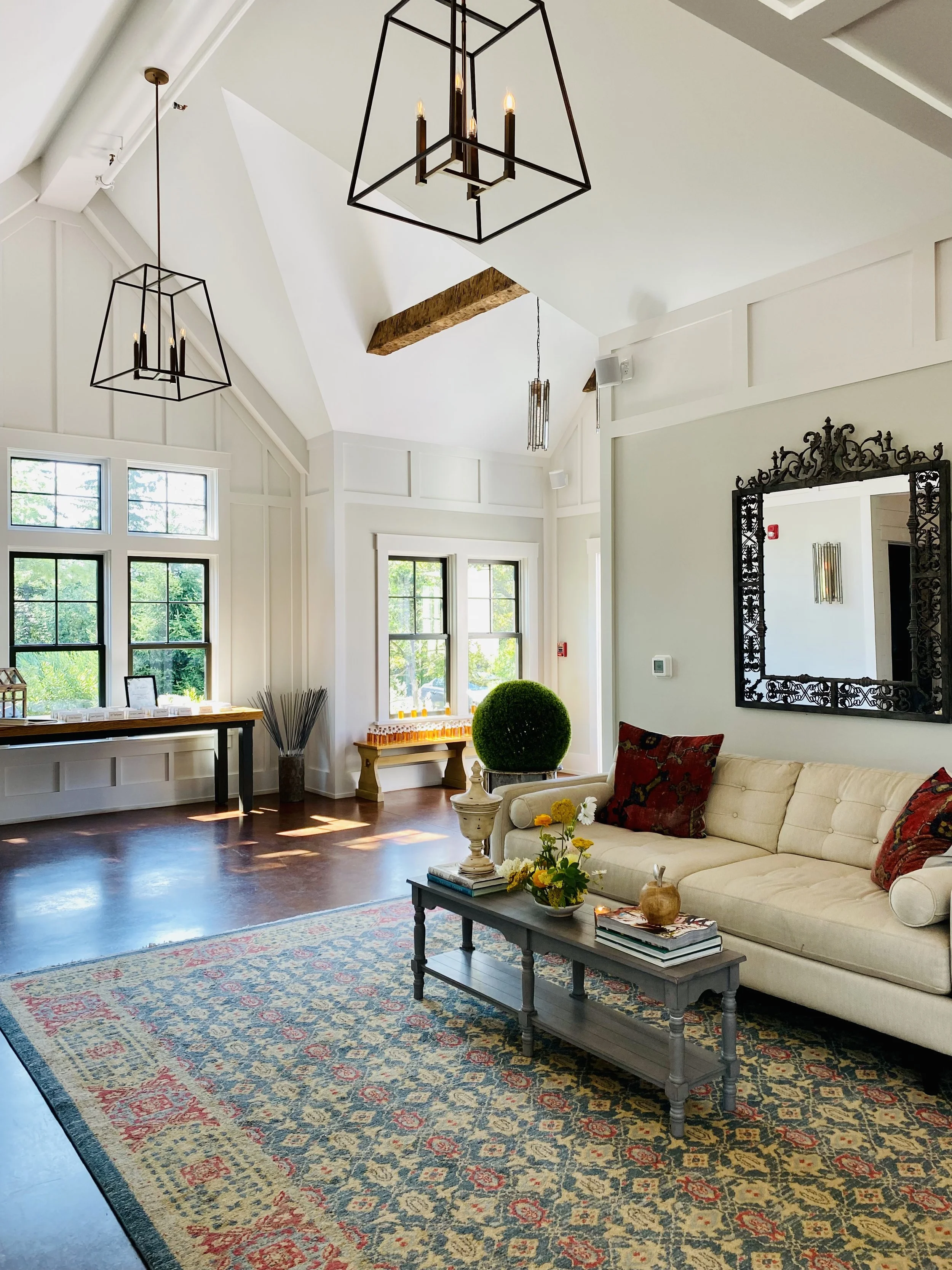 Interior of a bright, spacious living room with white walls, large windows, and dark wood flooring. Features include a beige sofa with patterned pillows, a black coffee table with books and decor, a floral area rug, and decorative elements like a topiary, candles, and framed art. Hanging geometric chandeliers and a large ornate mirror are visible.