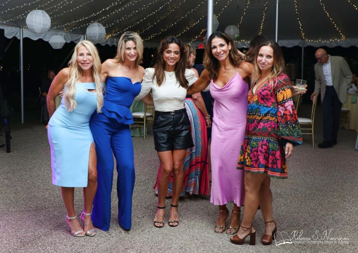 Five women standing together at a party under a tent decorated with string lights, dressed in colorful outfits, smiling at the camera.